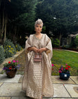 Woman in a flowing champagne gold Demure Boubou with matching headwrap, standing in a garden walkway with flowers.