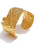 Hammered irregular metal cuff bracelets in gold and silver, displayed on a neutral surface with textured stone accents.