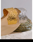 Hammered irregular metal cuff bracelets in gold and silver, displayed on a neutral surface with textured stone accents.