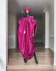Pink off-shoulder boubou dress with black grid pattern, paired with a matching headwrap, displayed on a mannequin against a black paneled wall.