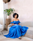 Model wearing a royal blue satin off-shoulder crop top and matching flared skirt in a minimalist living room setting.