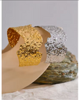 Hammered irregular metal cuff bracelets in gold and silver, displayed on a neutral surface with textured stone accents.