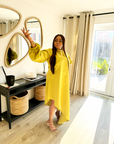A woman wearing a bright yellow satin slanted dress with long puff sleeves and a high-low hemline, posing indoors near a mirror and window.