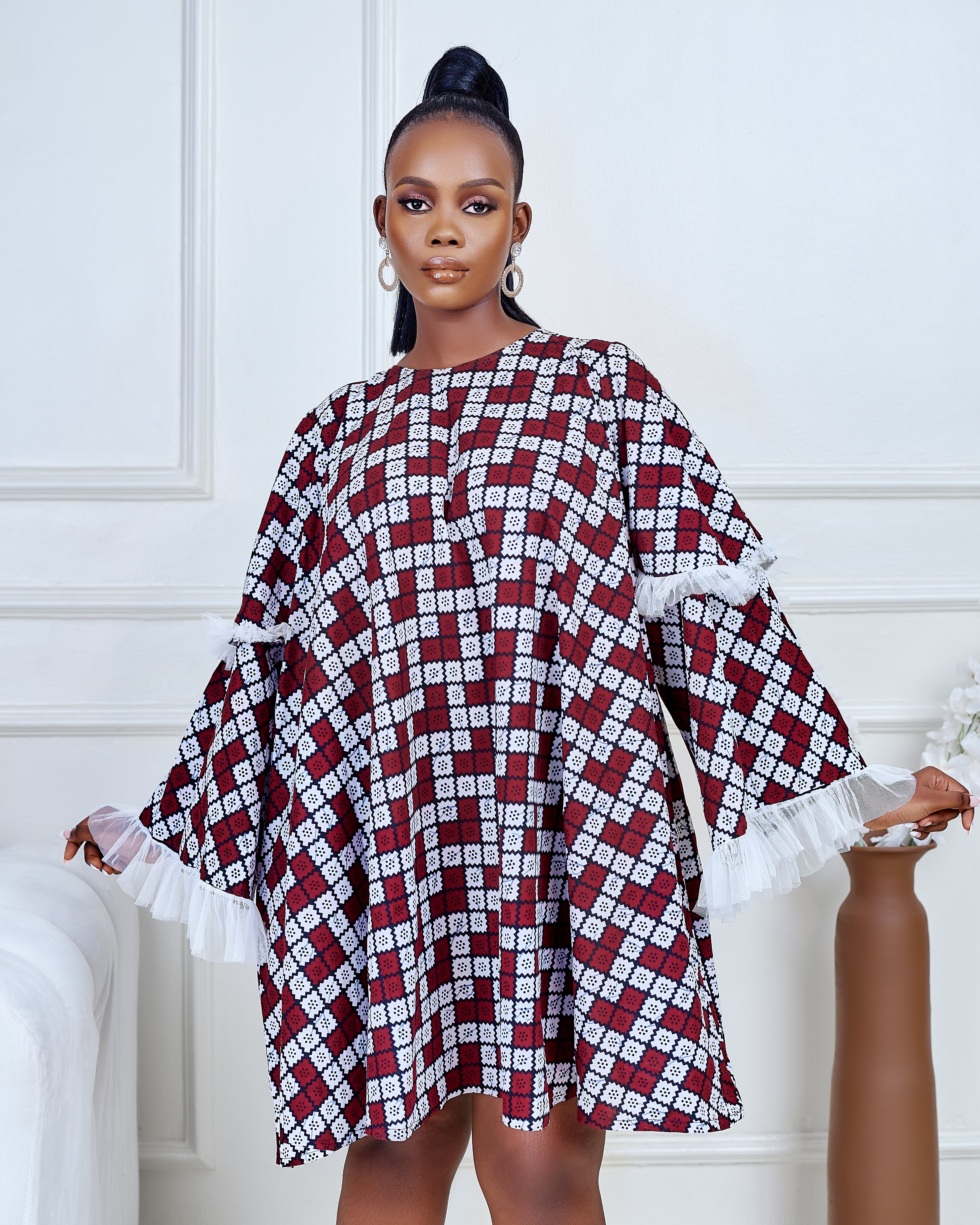 Model wearing a short flared Ankara dress with white lace sleeve detailing in a minimalist living room setting.
