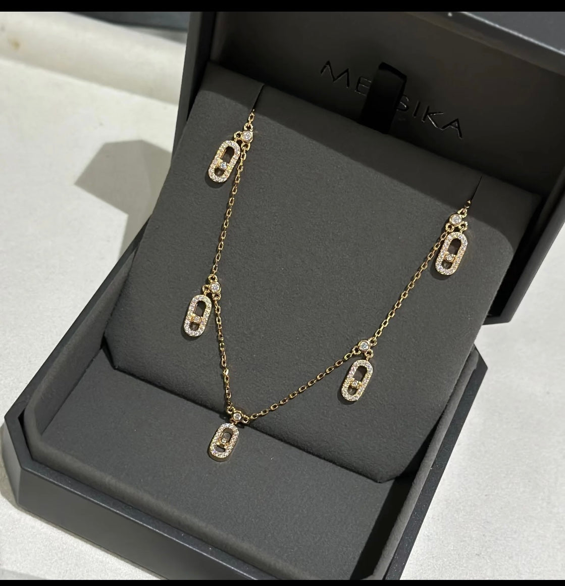 Gold chain necklace with crystal-studded oval pendants displayed in a black jewelry box.
