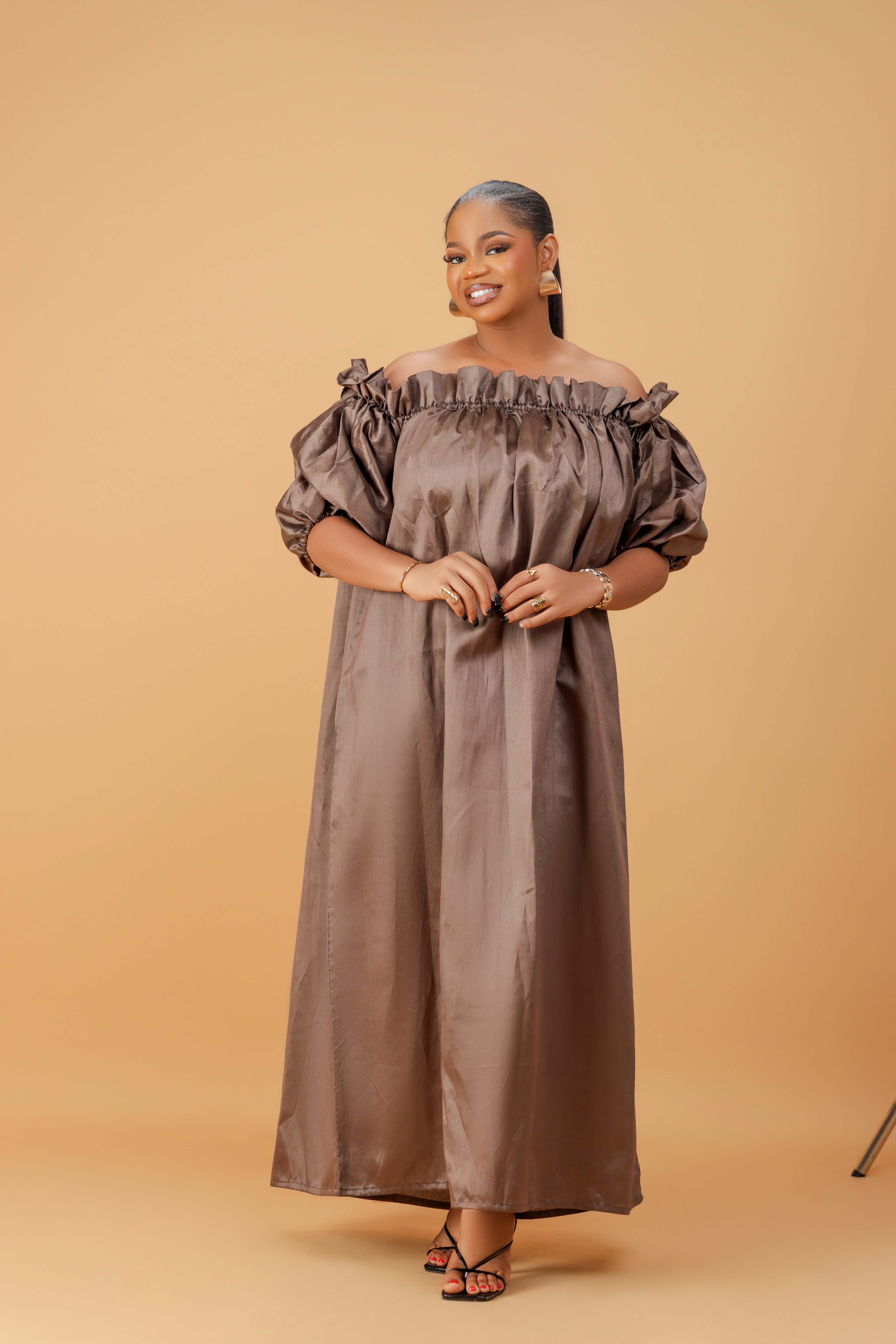 Woman wearing an off-shoulder brown maxi dress with puff sleeves and a flowing ankle-length design on a neutral background.

