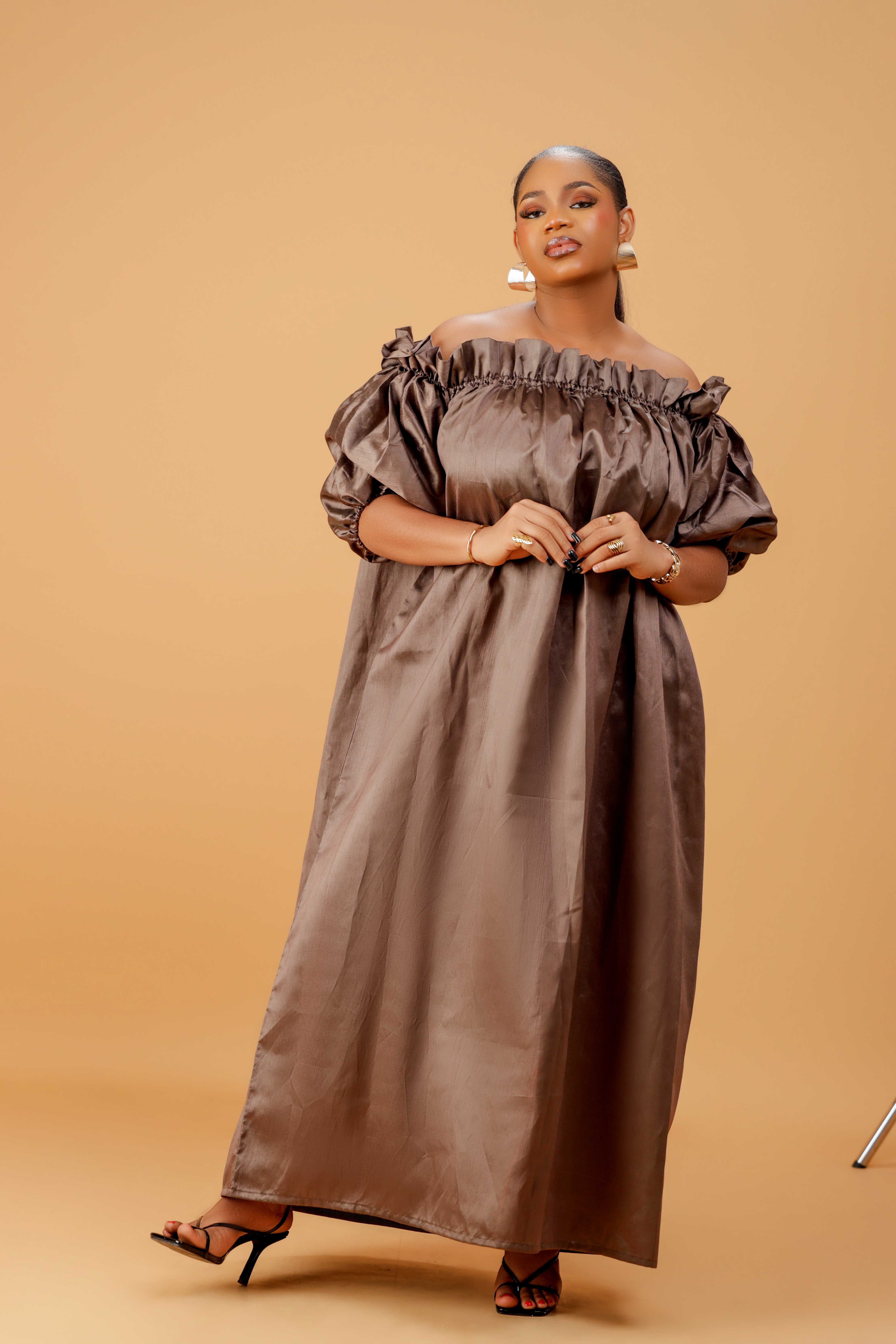 Woman wearing an off-shoulder brown maxi dress with puff sleeves and a flowing ankle-length design on a neutral background.
