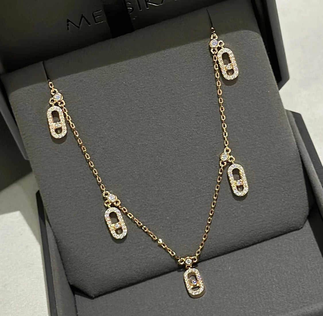 Gold chain necklace with crystal-studded oval pendants displayed in a black jewelry box.
