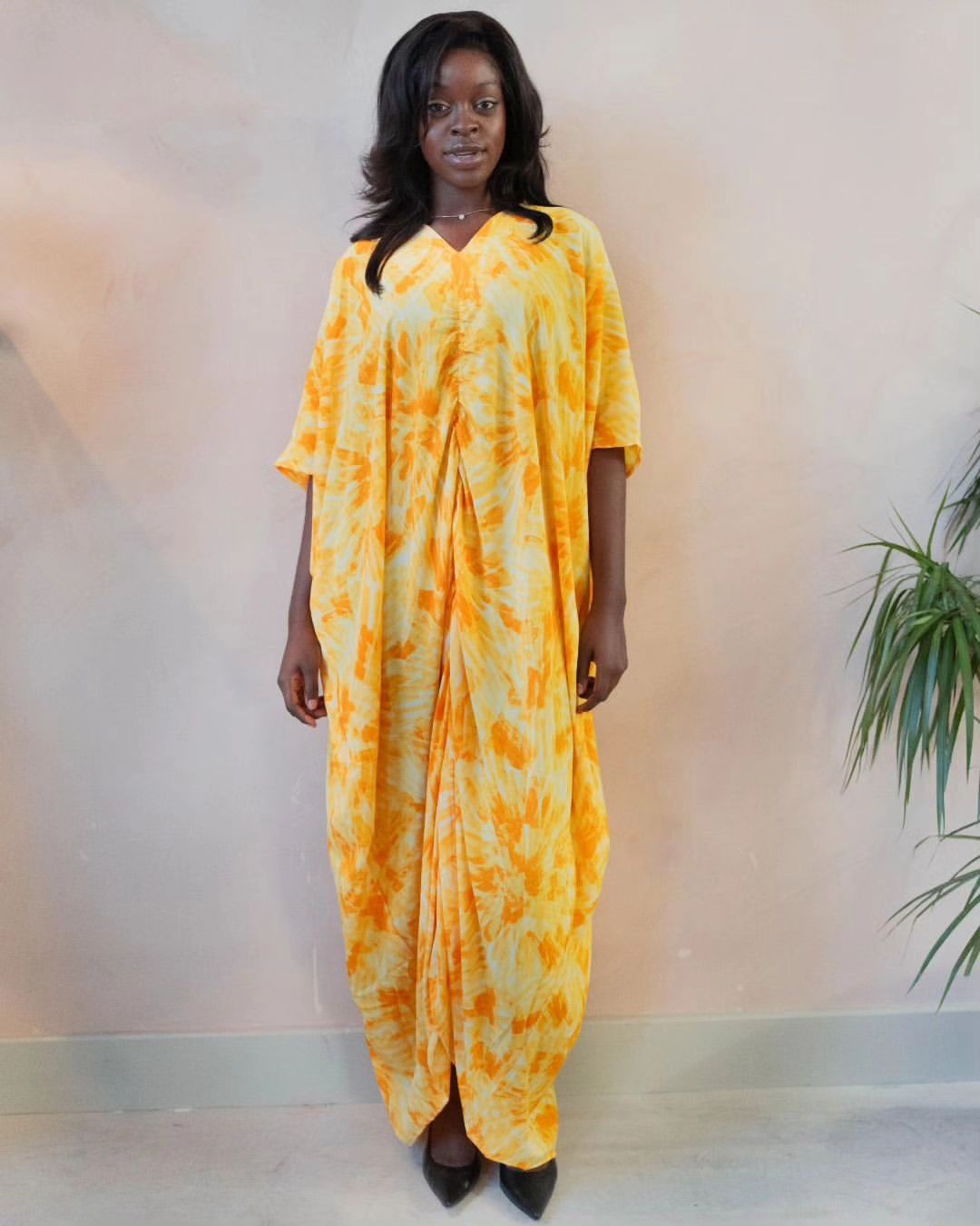 Woman wearing a yellow and orange tie-dye draped maxi dress with front gathers, standing indoors.
