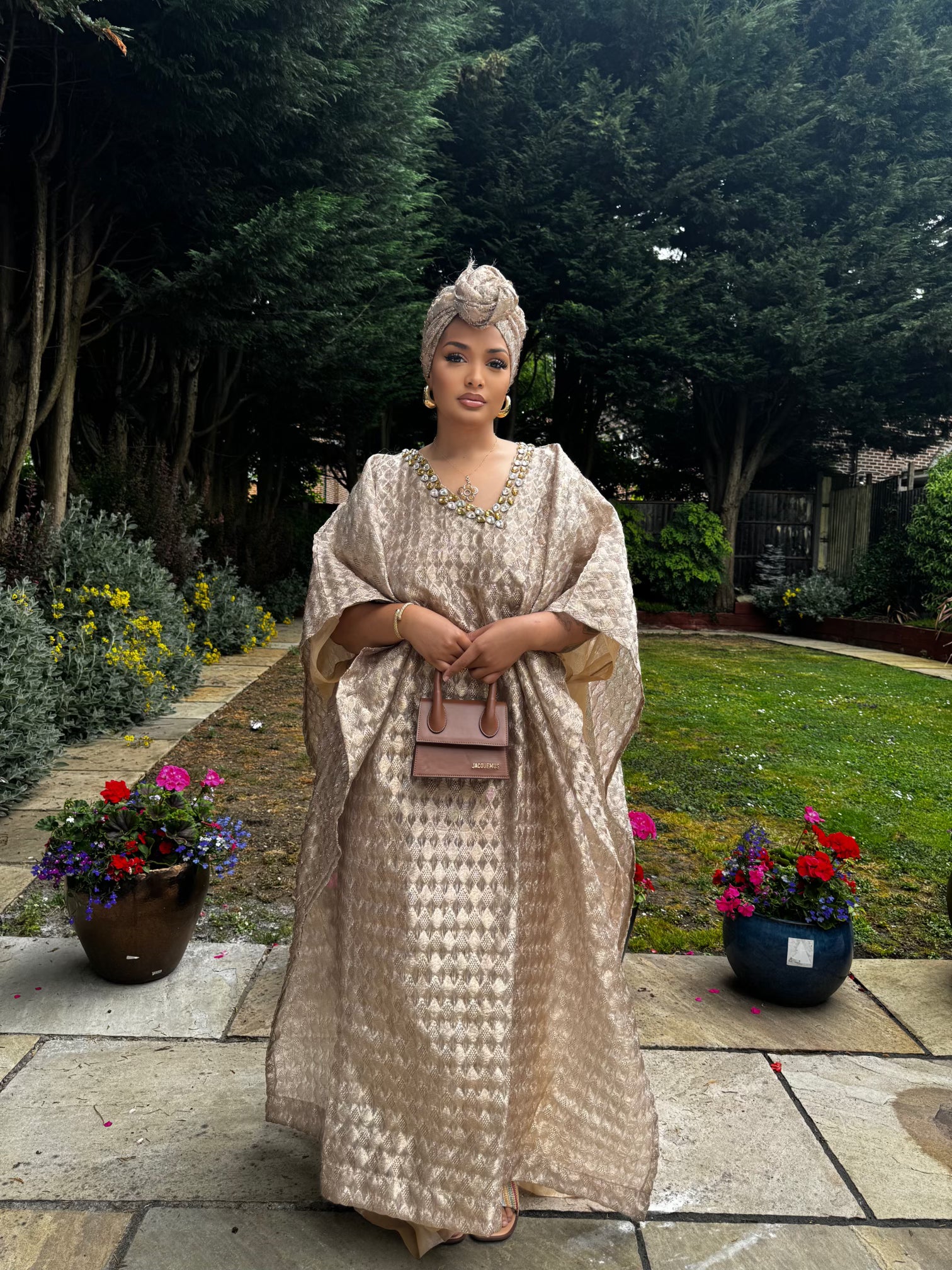  Woman in a flowing champagne gold Demure Boubou with matching headwrap, standing in a garden walkway with flowers.
