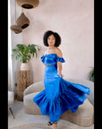 Model wearing a royal blue satin off-shoulder crop top and matching flared skirt in a minimalist living room setting.
