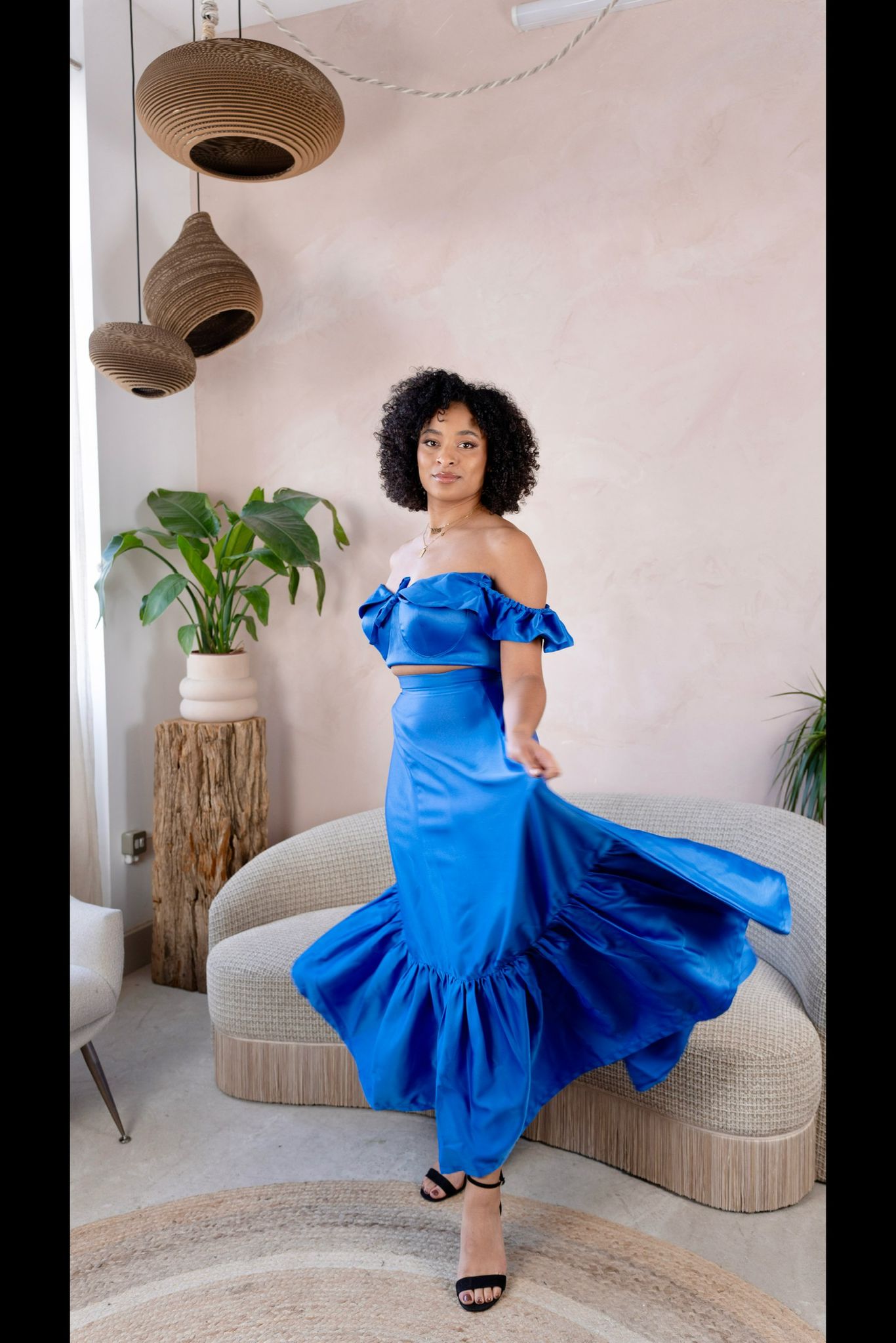 Model wearing a royal blue satin off-shoulder crop top and matching flared skirt in a minimalist living room setting.

