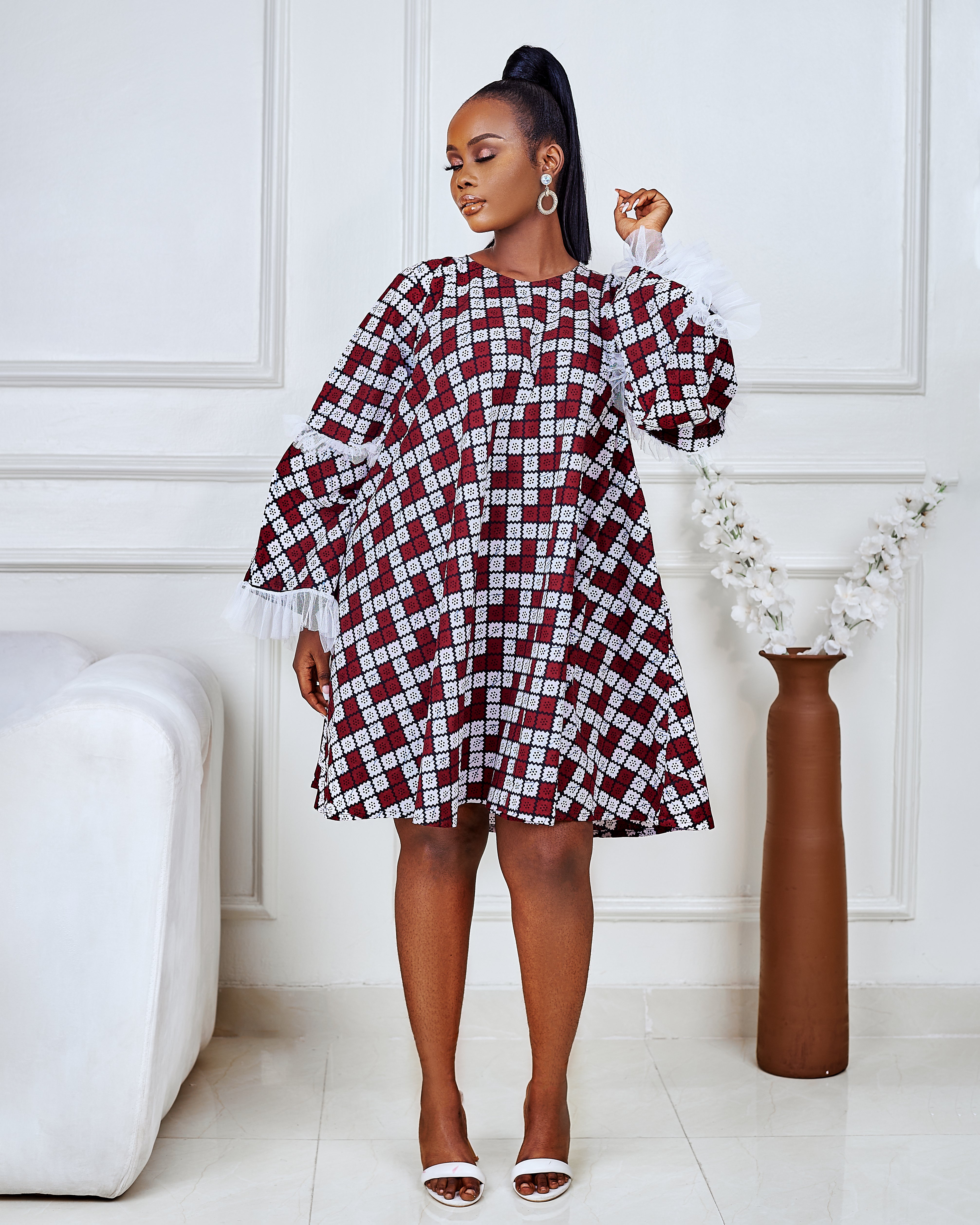 Model wearing a short flared Ankara dress with white lace sleeve detailing in a minimalist living room setting.
