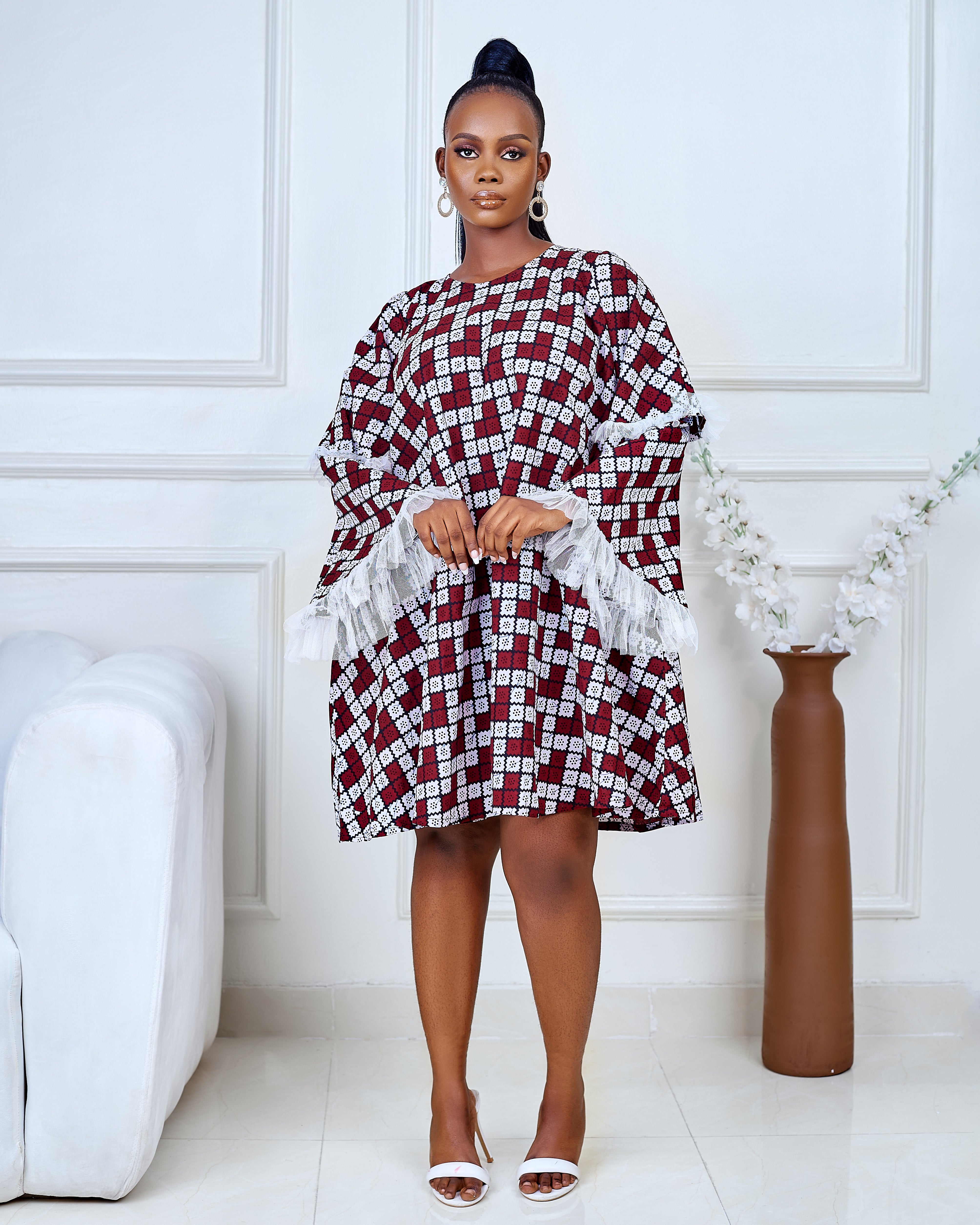 Model wearing a short flared Ankara dress with white lace sleeve detailing in a minimalist living room setting.
