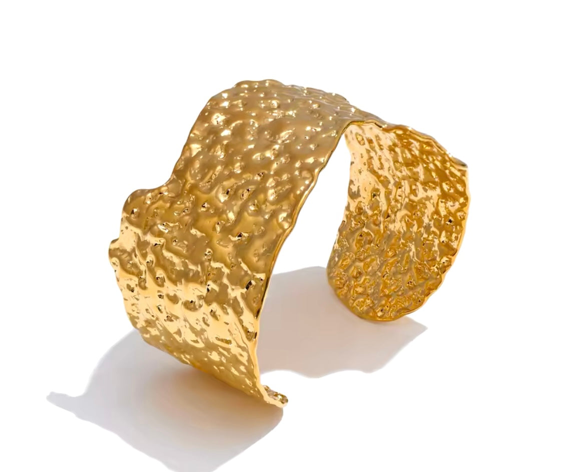 Hammered irregular metal cuff bracelets in gold and silver, displayed on a neutral surface with textured stone accents.

