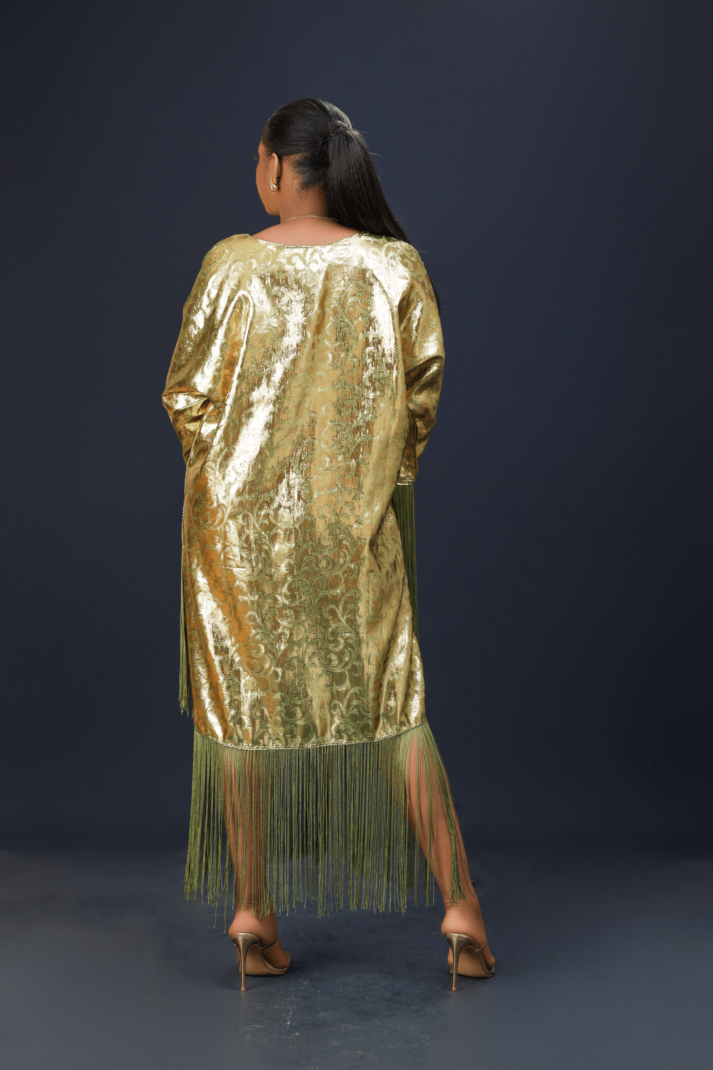 A woman wearing a shimmering gold kaftan dress with long green fringes, standing against a dark background.
