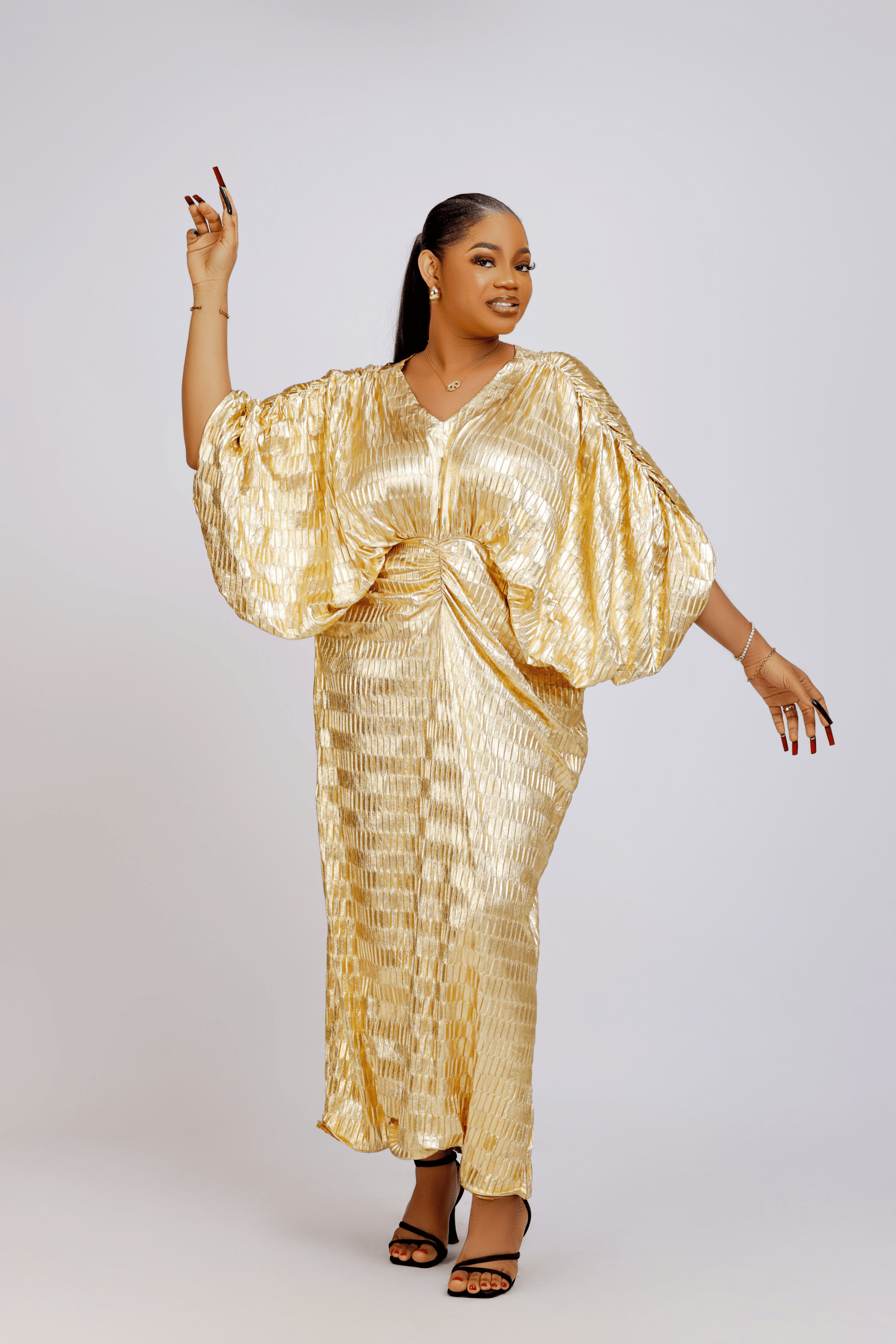 Woman wearing a gold metallic draped dress with voluminous sleeves and a cinched waist, posing against a light background.
