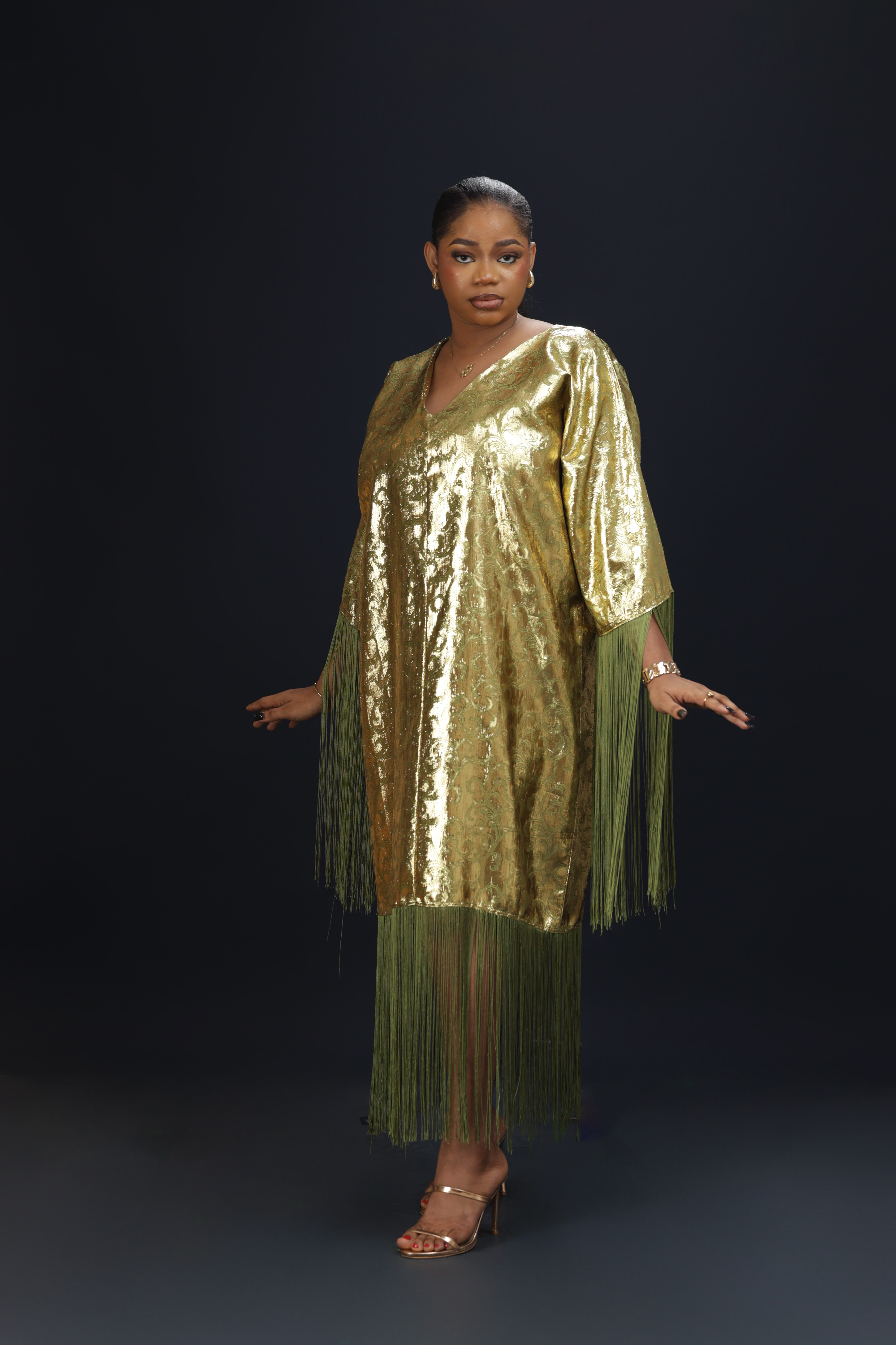 A woman wearing a shimmering gold kaftan dress with long green fringes, standing against a dark background.
