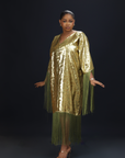 A woman wearing a shimmering gold kaftan dress with long green fringes, standing against a dark background.

