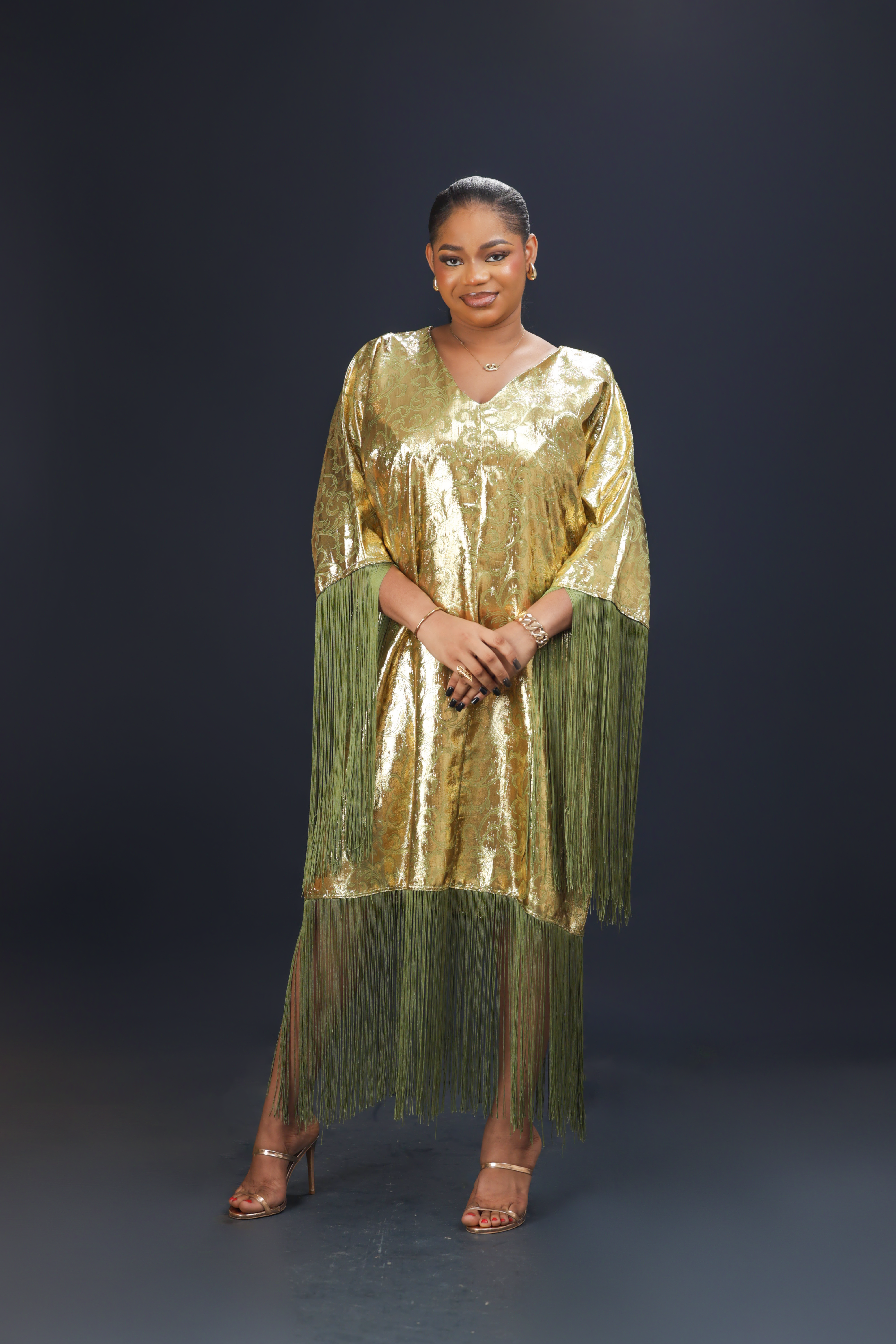 A woman wearing a shimmering gold kaftan dress with long green fringes, standing against a dark background.
