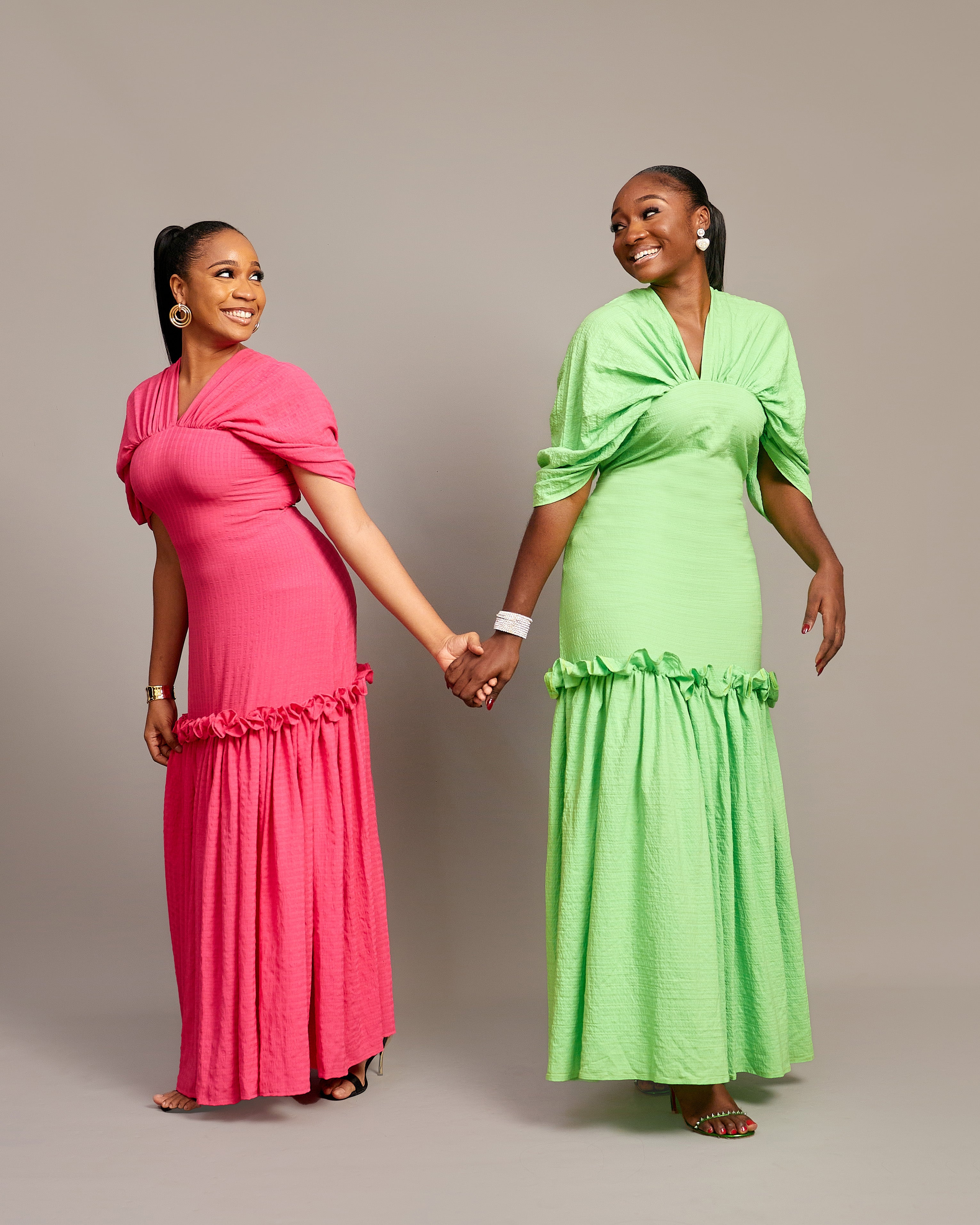 Two women wearing the Ine maxi dress in pink and green, featuring a draped V neckline, gathered waist detailing, and ruched hem accents.
