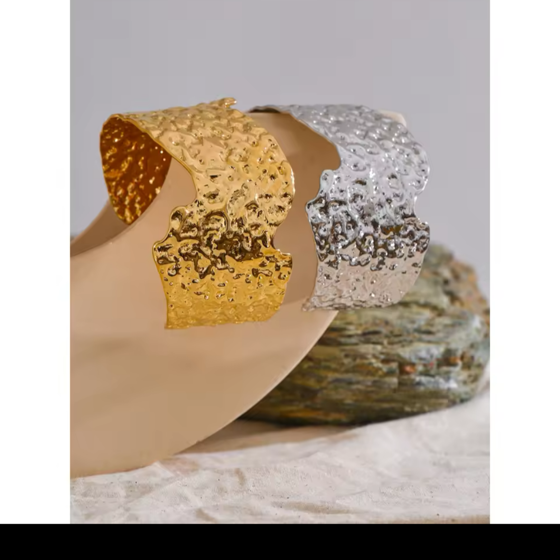 Hammered irregular metal cuff bracelets in gold and silver, displayed on a neutral surface with textured stone accents.
