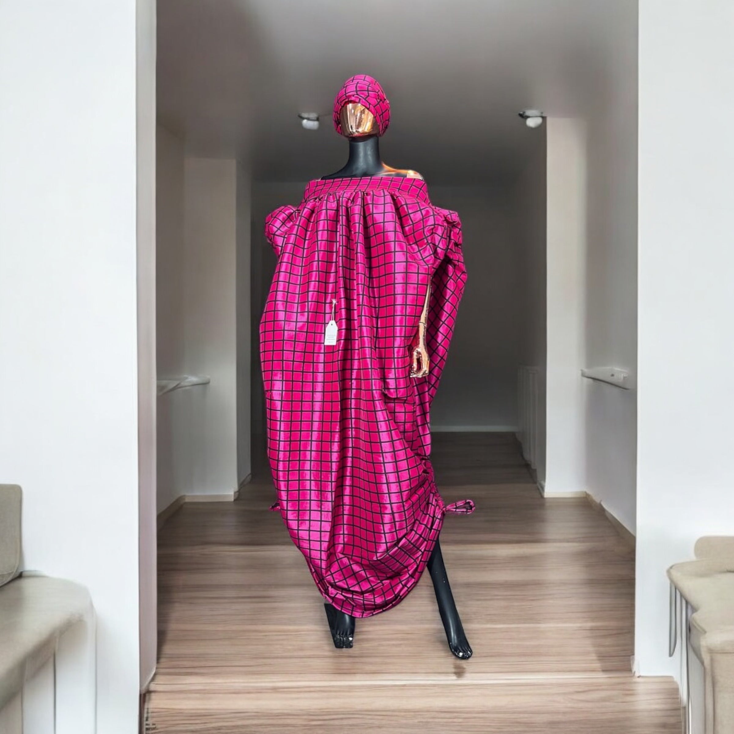 Pink off-shoulder boubou dress with black grid pattern, paired with a matching headwrap, displayed on a mannequin against a black paneled wall.
