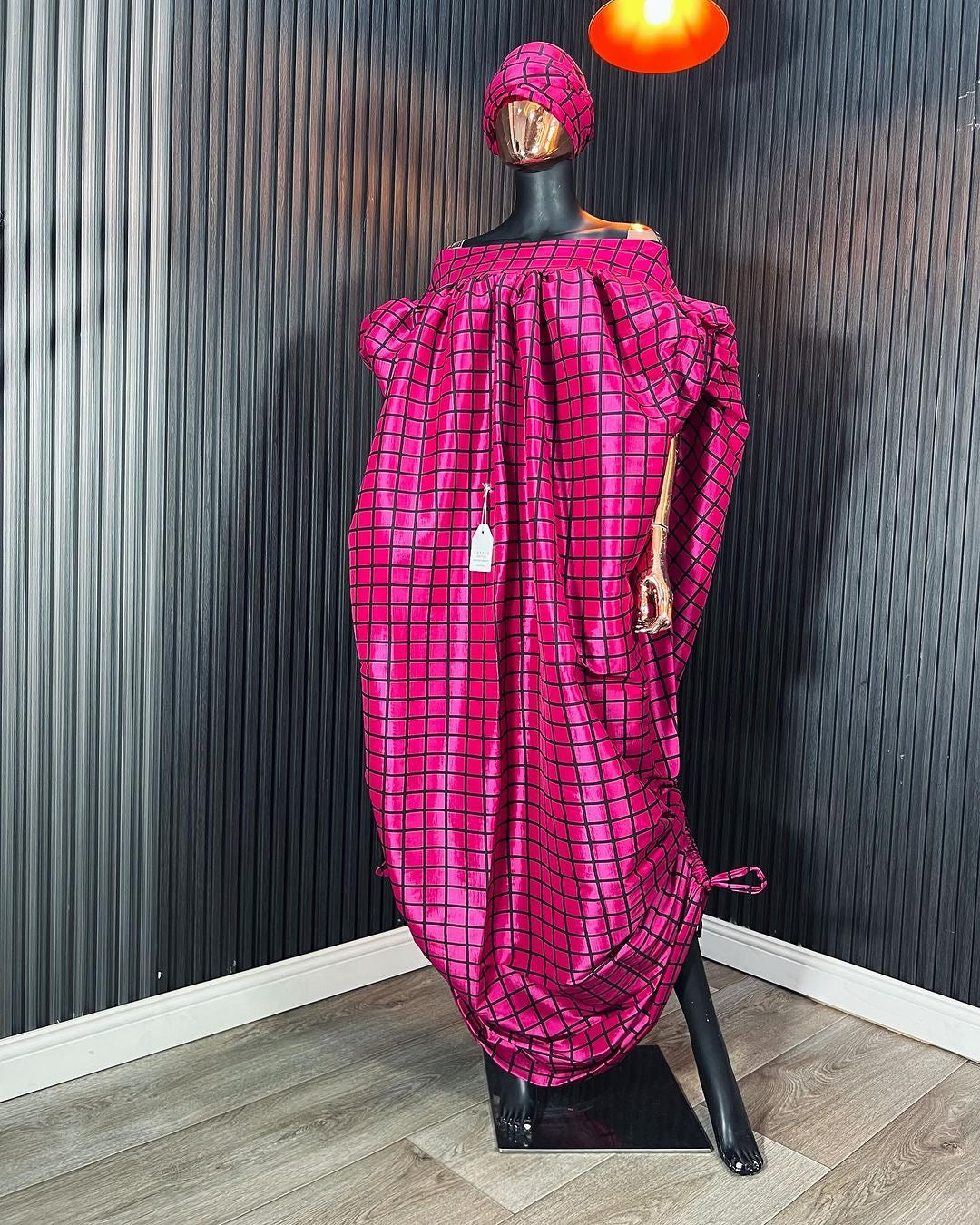 Pink off-shoulder boubou dress with black grid pattern, paired with a matching headwrap, displayed on a mannequin against a black paneled wall.
