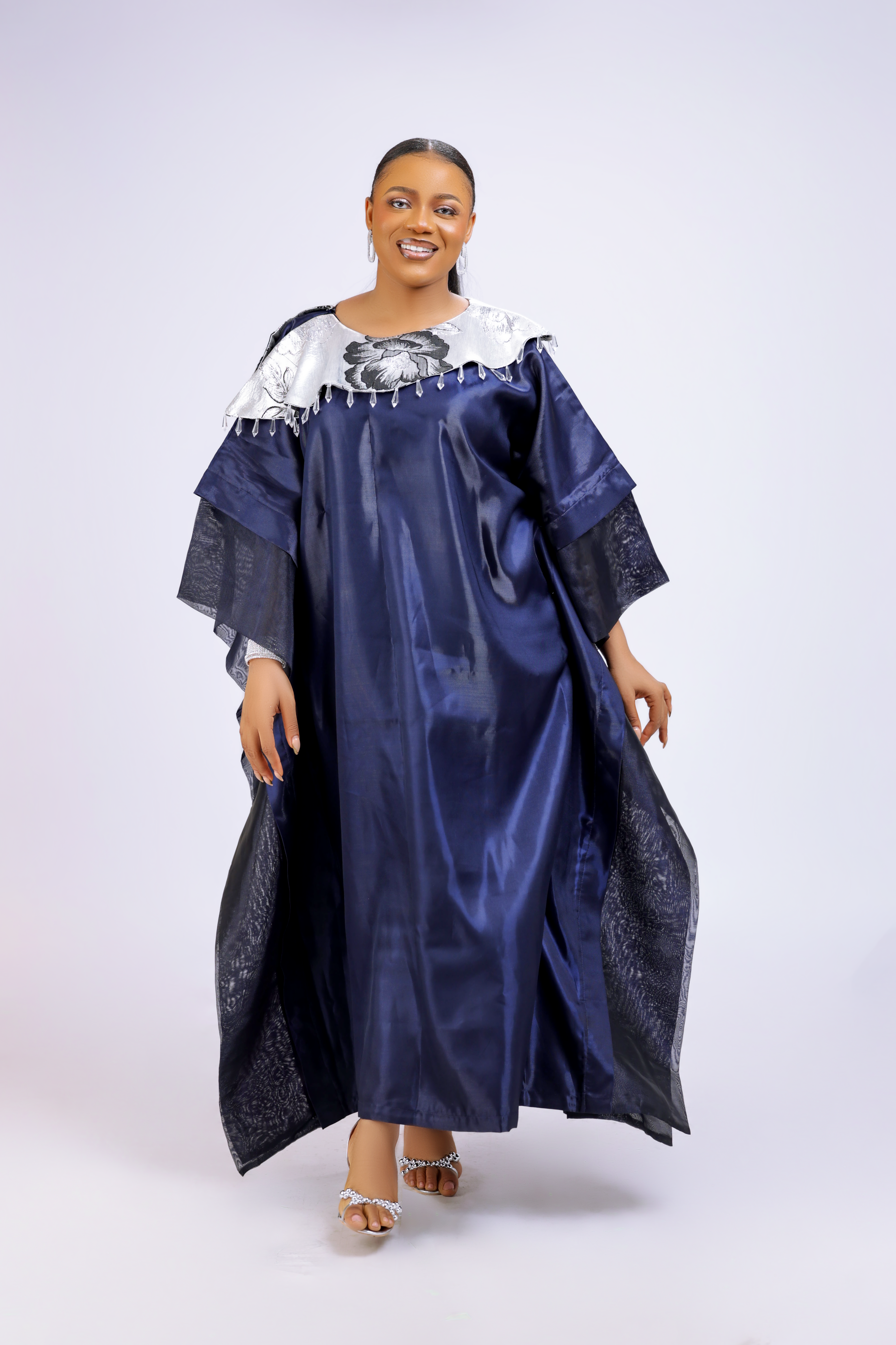 A woman wearing a metallic navy blue kaftan gown with an embroidered white floral neckline and wide sleeves, styled for special occasions.
