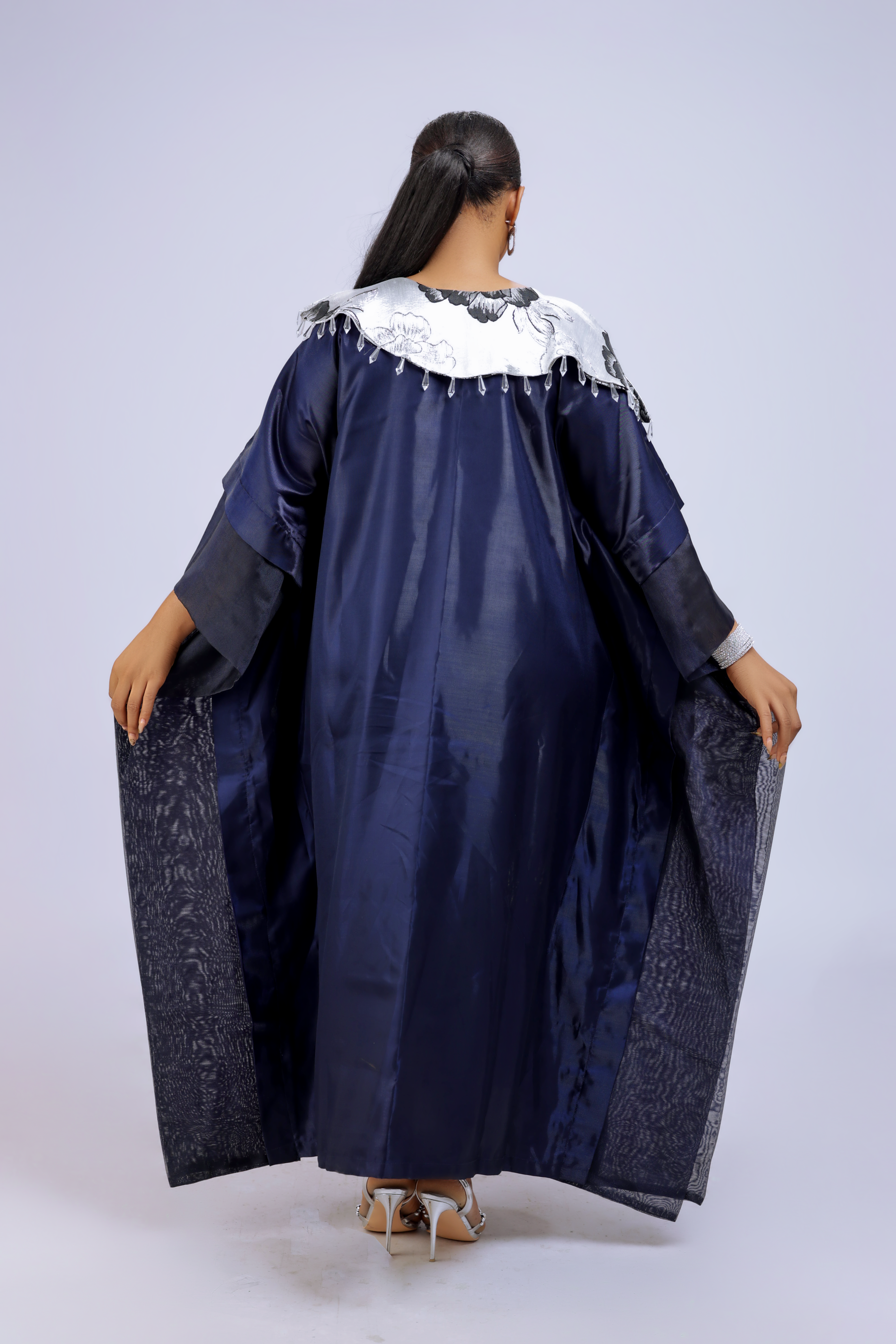 A woman wearing a metallic navy blue kaftan gown with an embroidered white floral neckline and wide sleeves, styled for special occasions.
