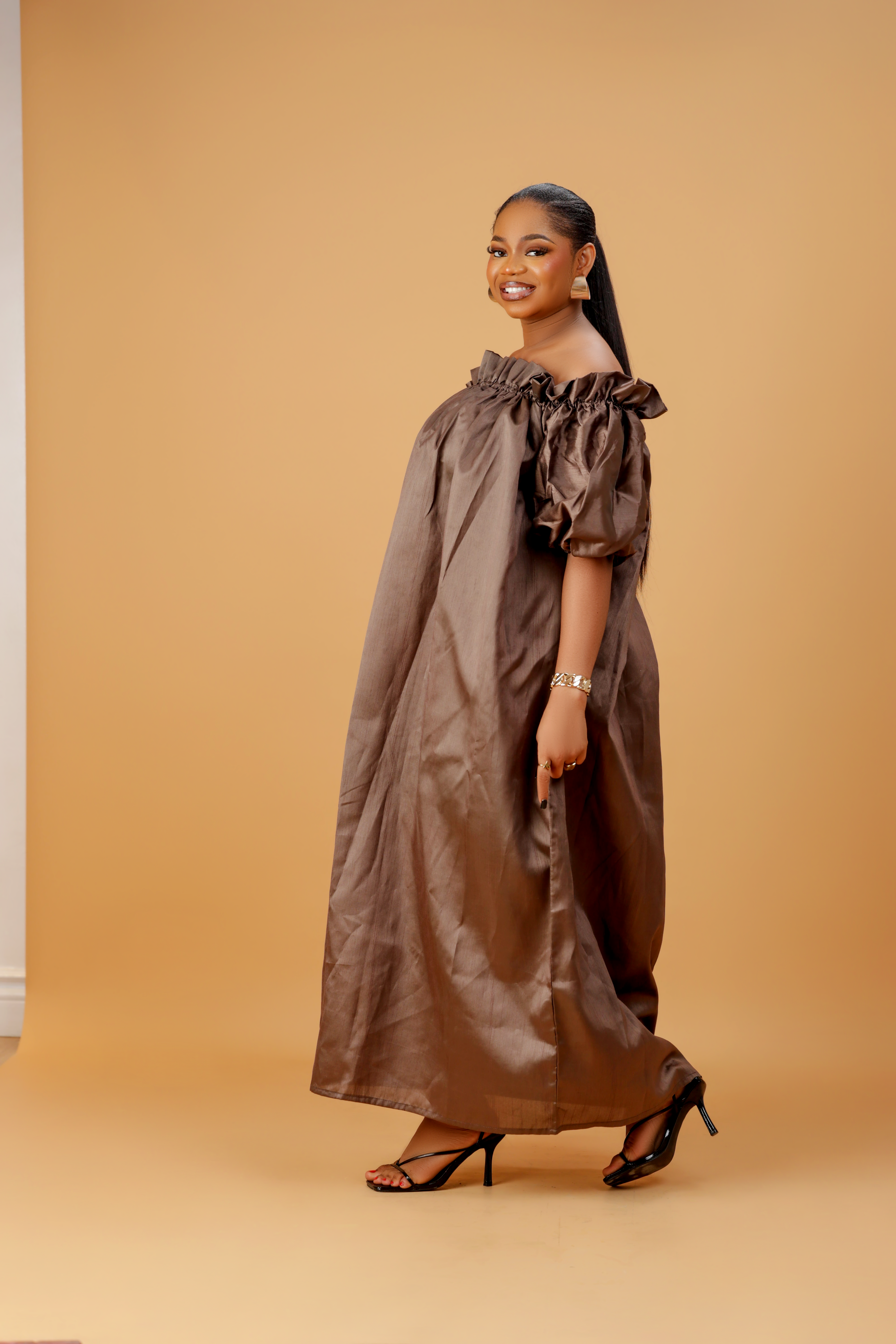 Woman wearing an off-shoulder brown maxi dress with puff sleeves and a flowing ankle-length design on a neutral background.
