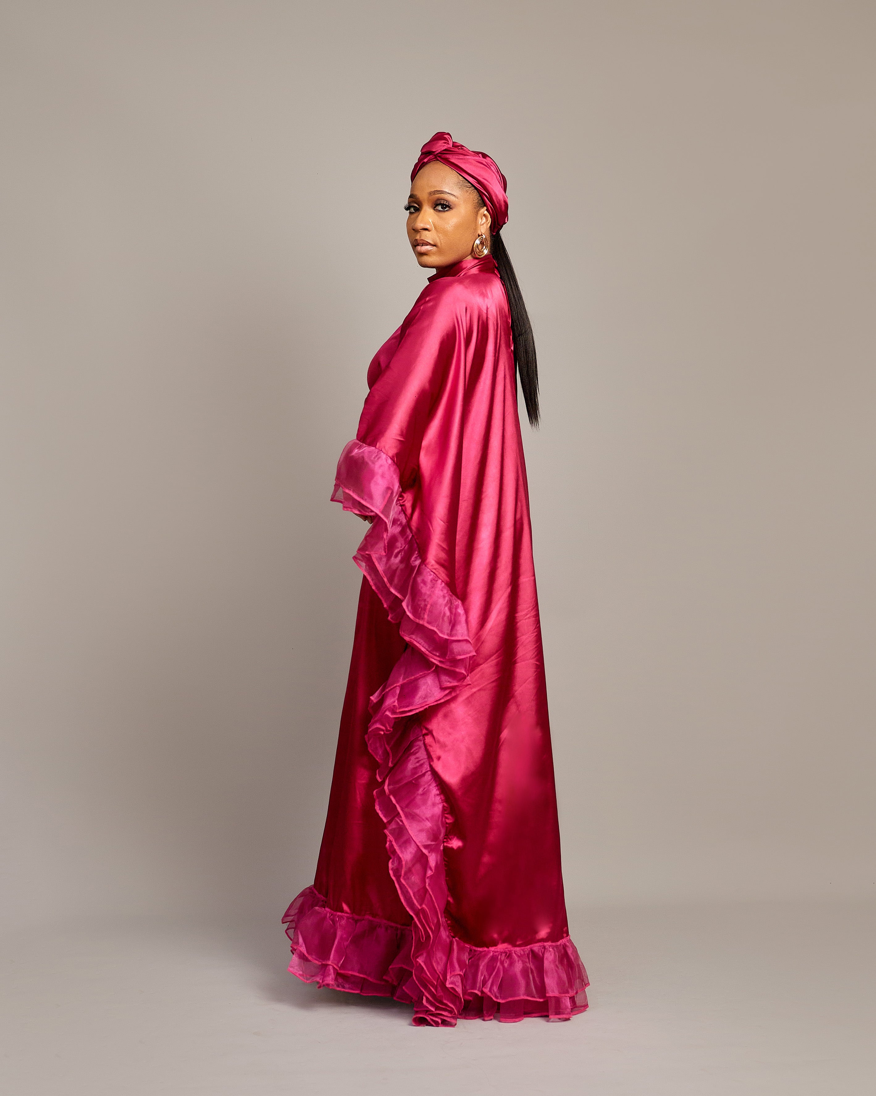Woman wearing the Ola Buubu satin gown in pink with ruffle edges, long sleeves, high neckline, and matching headwrap.
