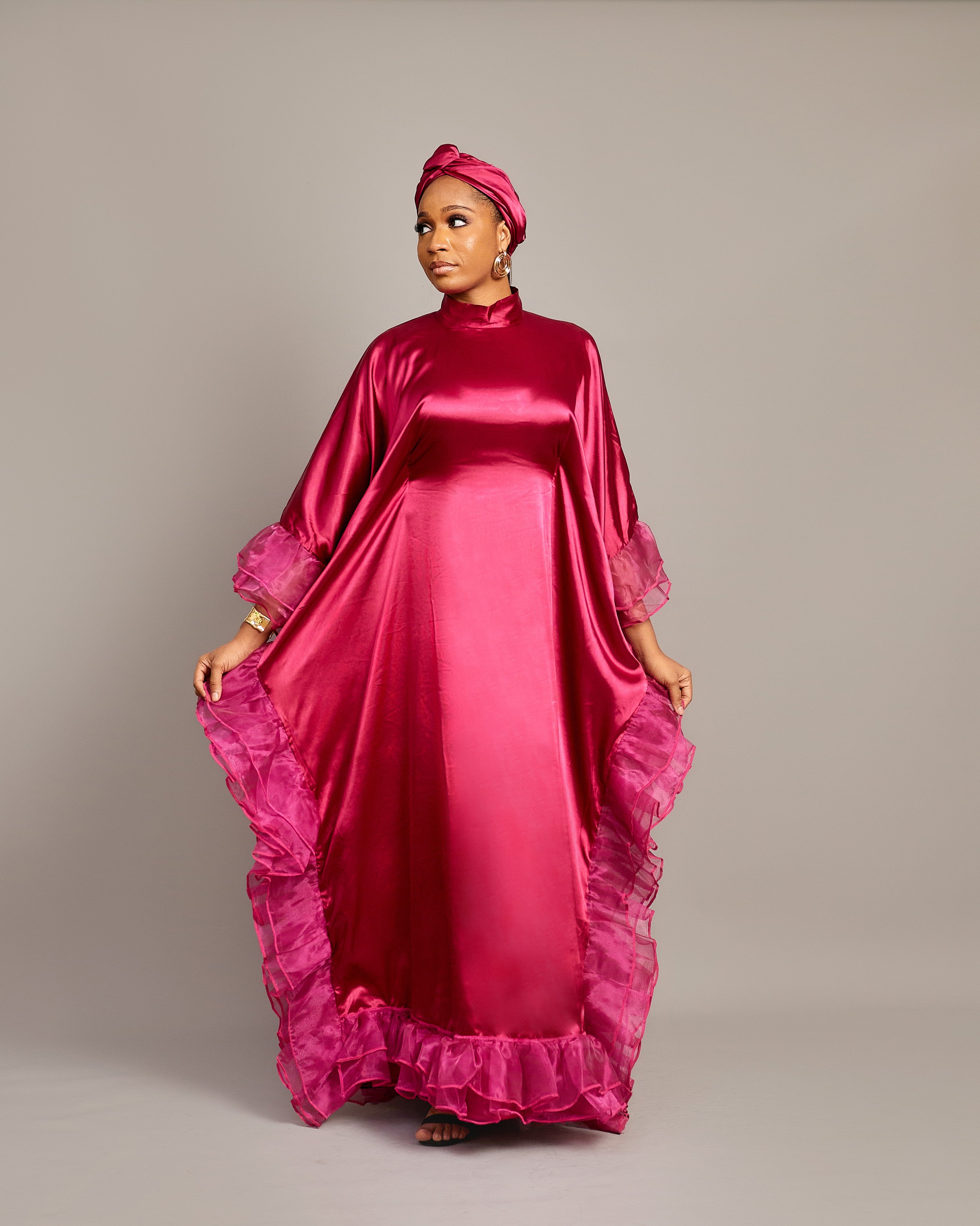 Woman wearing the Ola Buubu satin gown in pink with ruffle edges, long sleeves, high neckline, and matching headwrap.
