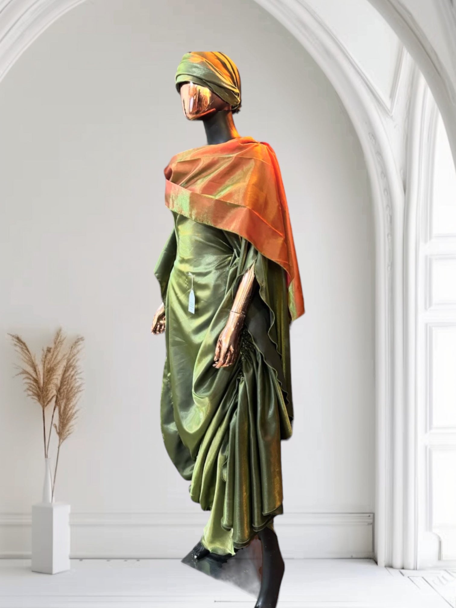 Olive green satin gown with ruched side draping, paired with a bronze-toned shawl and matching headwrap, displayed on a mannequin in an arched interior.

