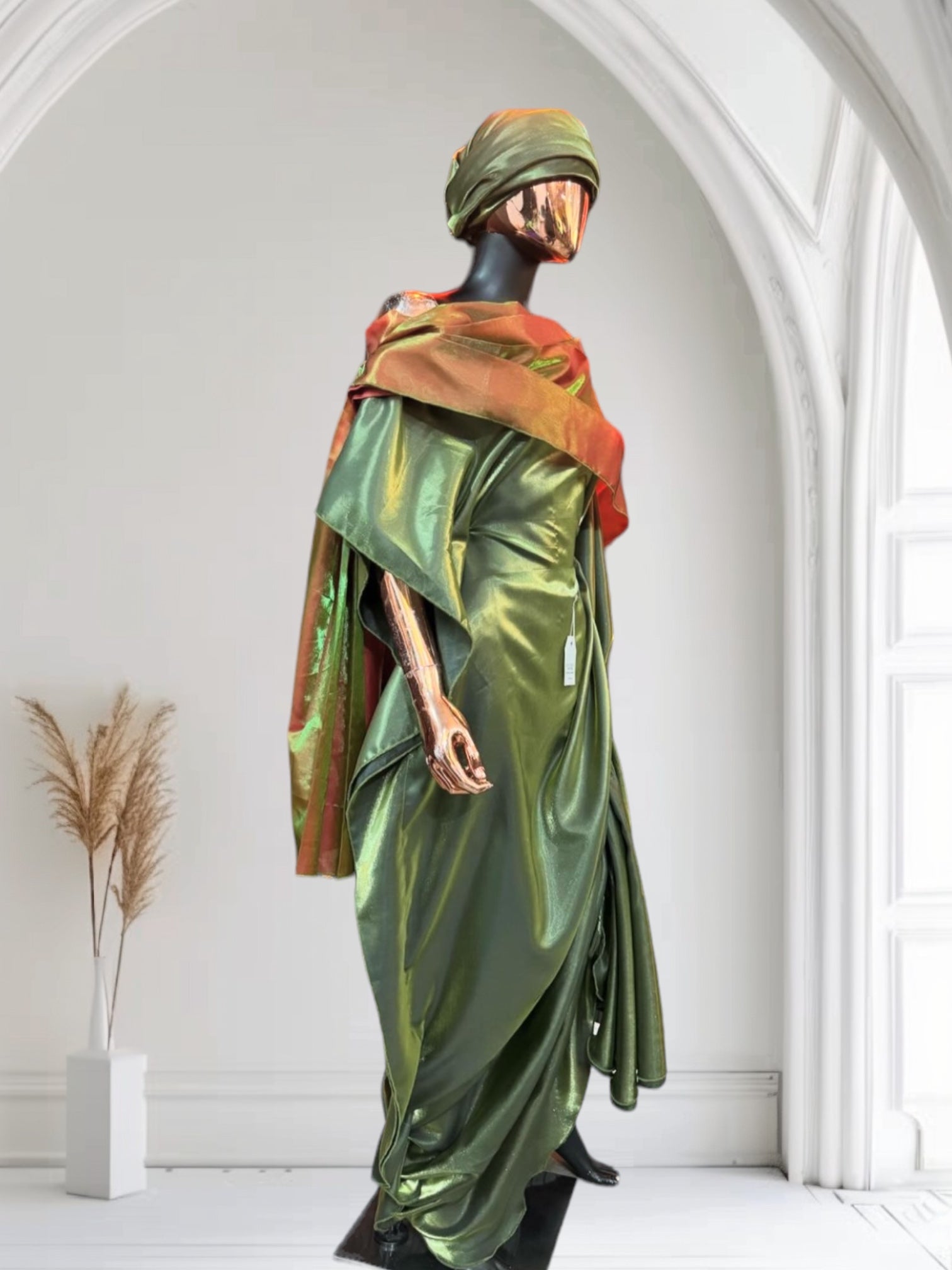 Olive green satin gown with ruched side draping, paired with a bronze-toned shawl and matching headwrap, displayed on a mannequin in an arched interior.
