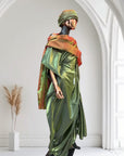 Olive green satin gown with ruched side draping, paired with a bronze-toned shawl and matching headwrap, displayed on a mannequin in an arched interior.
