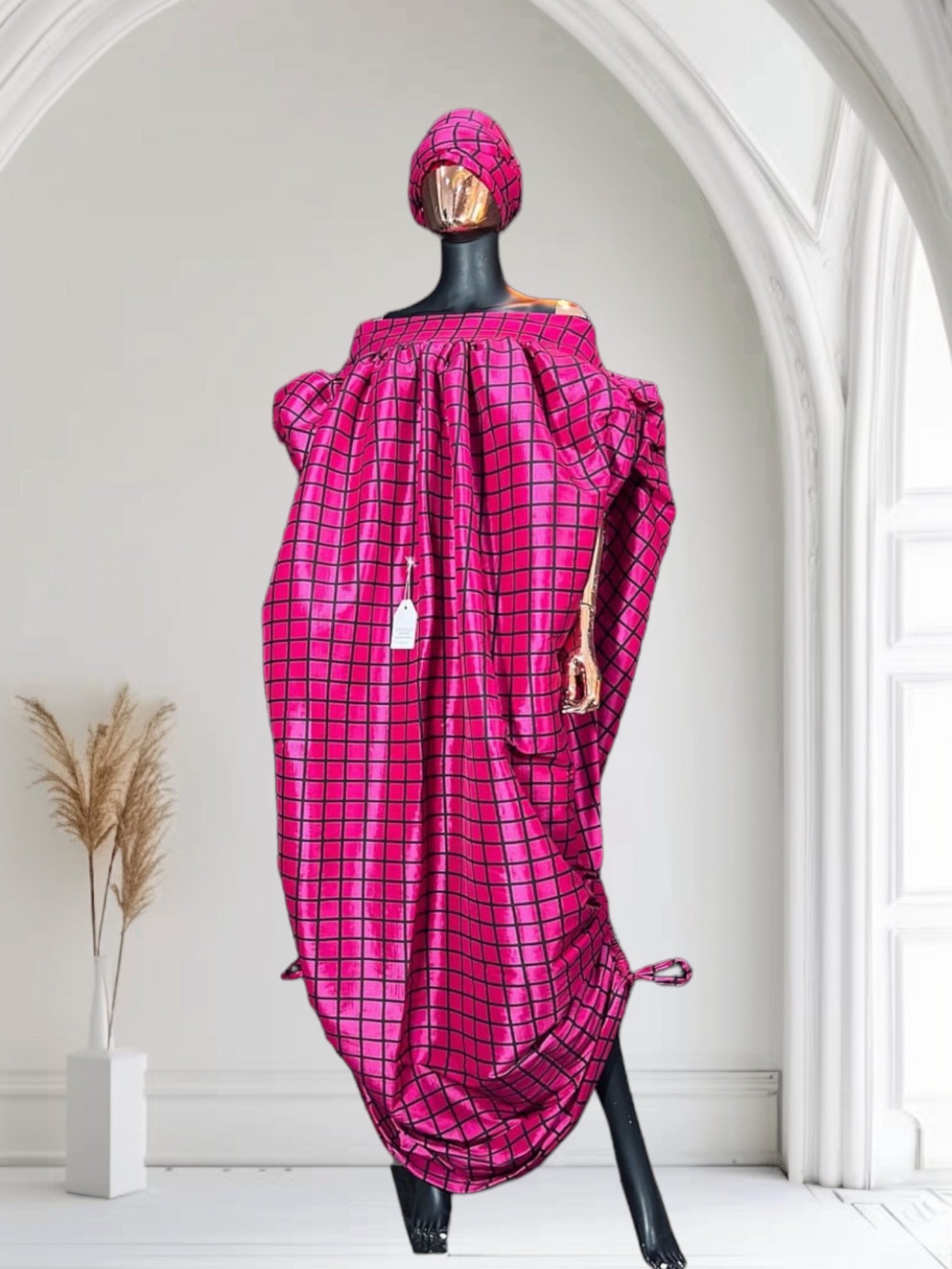 Pink off-shoulder boubou dress with black grid pattern, paired with a matching headwrap, displayed on a mannequin against a black paneled wall.
