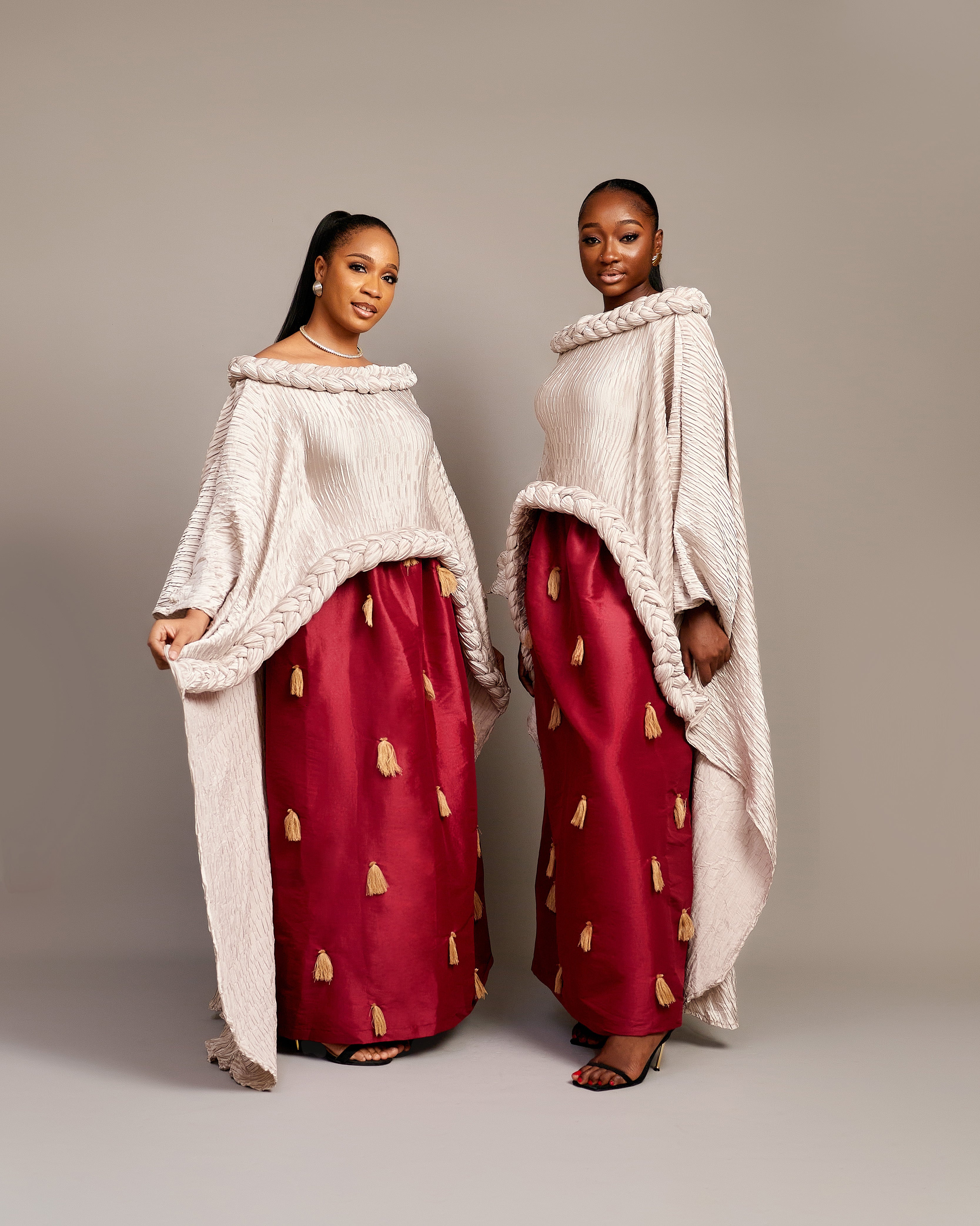 Two women wearing the Queen Ire two-piece set with textured ivory cape tops and wine maxi skirts decorated with golden tassels, styled for weddings and cultural celebrations.
