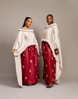 Two women wearing the Queen Ire two-piece set with textured ivory cape tops and wine maxi skirts decorated with golden tassels, styled for weddings and cultural celebrations.
