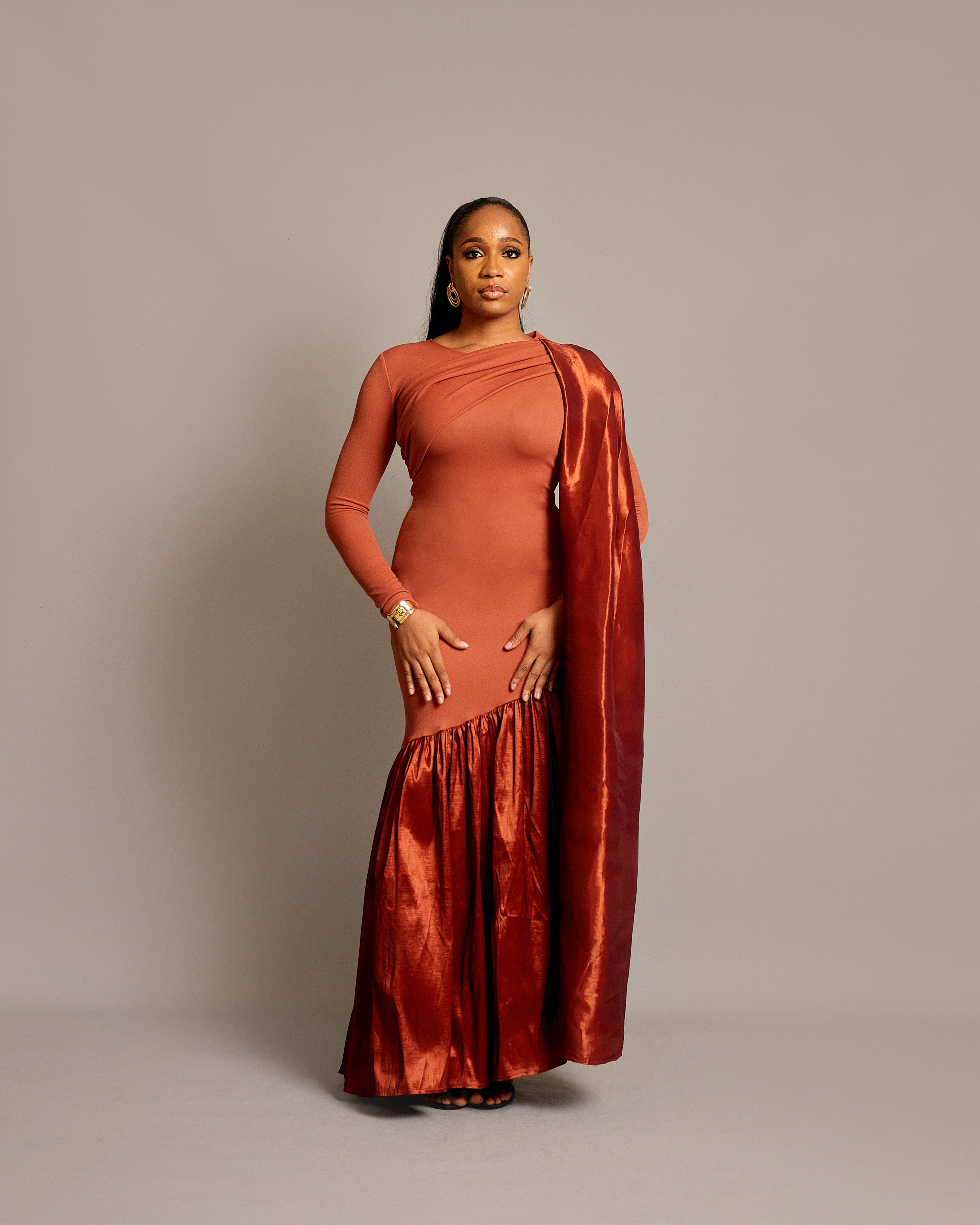 A woman wearing a rust sari dress with an asymmetrical neckline, gathered satin hem, and draped fabric over one shoulder.
