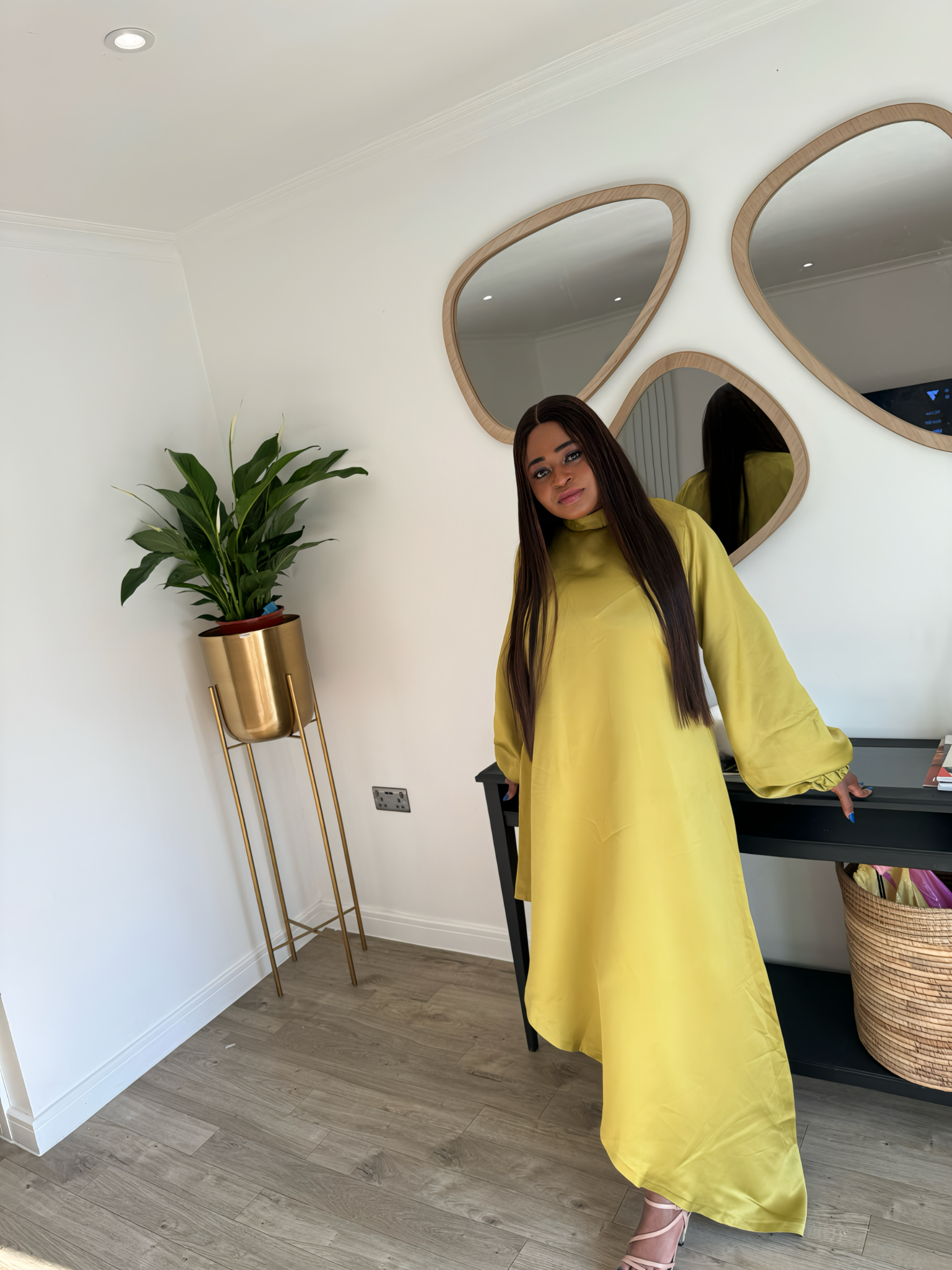 A woman wearing a bright yellow satin slanted dress with long puff sleeves and a high-low hemline, posing indoors near a mirror and window.
