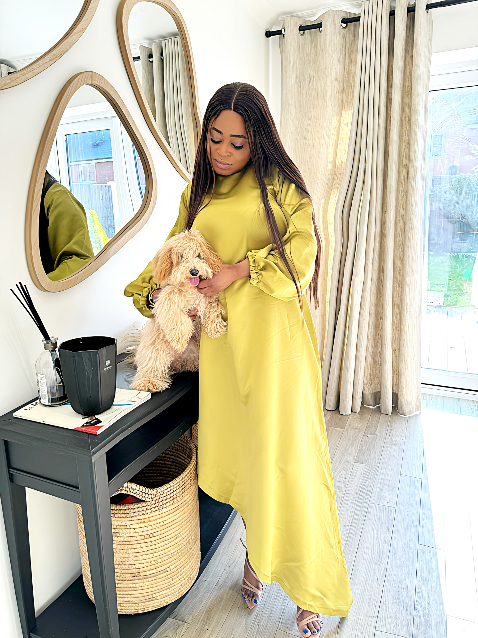 A woman wearing a bright yellow satin slanted dress with long puff sleeves and a high-low hemline, posing indoors near a mirror and window.
