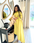A woman wearing a bright yellow satin slanted dress with long puff sleeves and a high-low hemline, posing indoors near a mirror and window.
