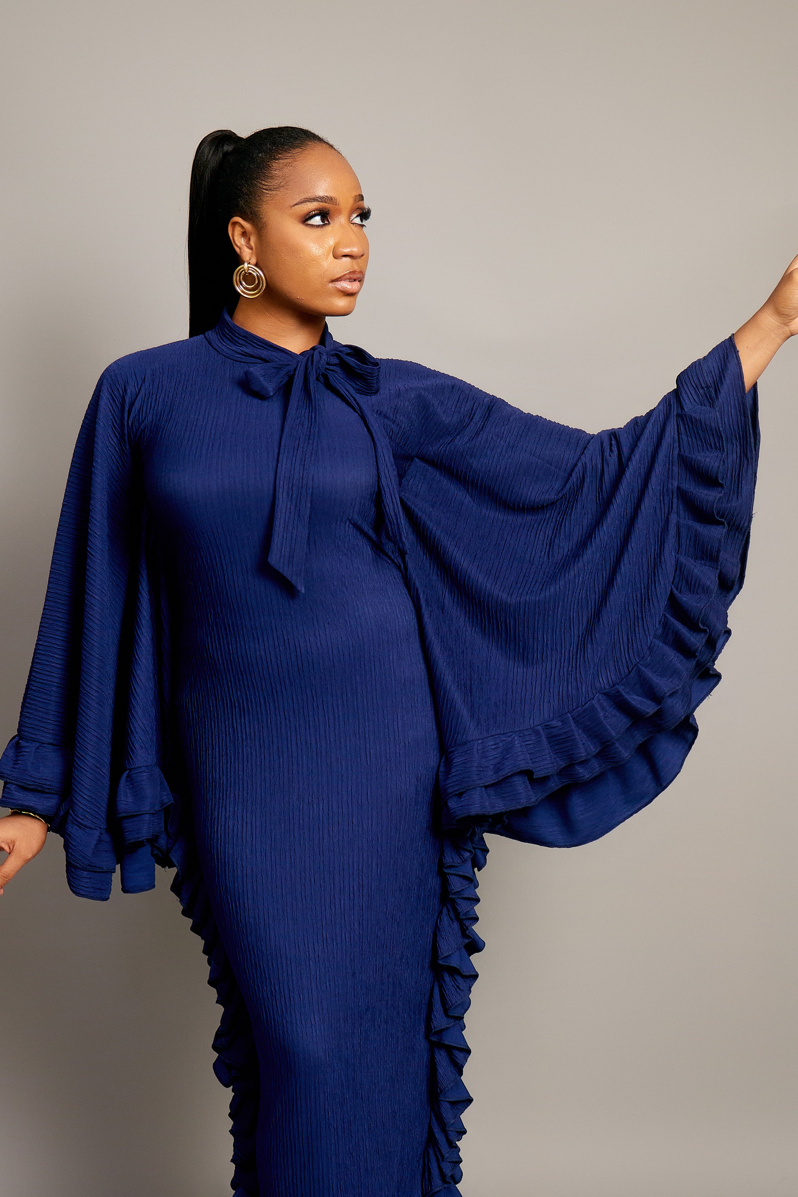 woman wearing the Tella Dress: one in ivory, one in navy. Maxi gowns with ruffle sides, long sleeves, and bow-tie neckline.
