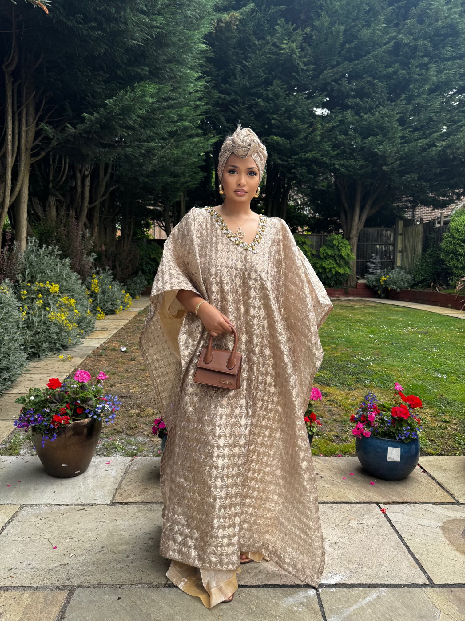  Woman in a flowing champagne gold Demure Boubou with matching headwrap, standing in a garden walkway with flowers.
