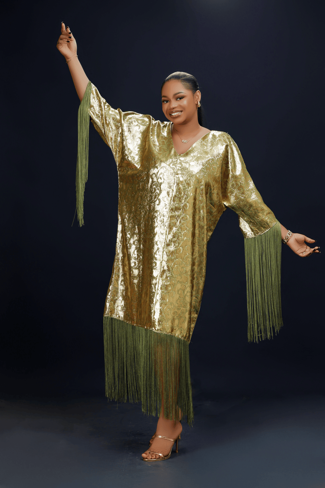 A woman wearing a shimmering gold kaftan dress with long green fringes, standing against a dark background.
