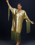 A woman wearing a shimmering gold kaftan dress with long green fringes, standing against a dark background.
