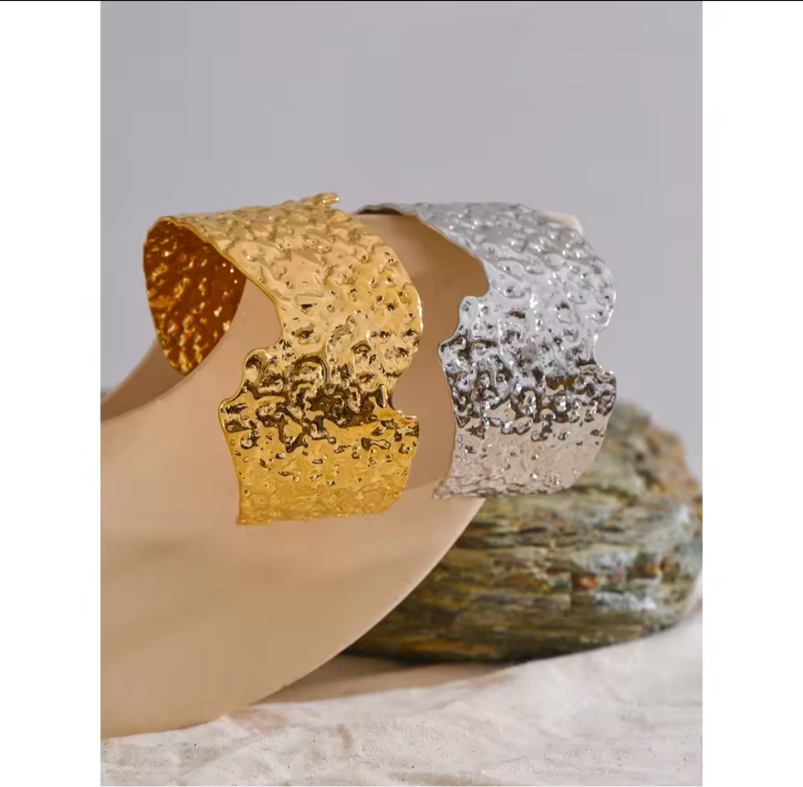 Hammered irregular metal cuff bracelets in gold and silver, displayed on a neutral surface with textured stone accents.
