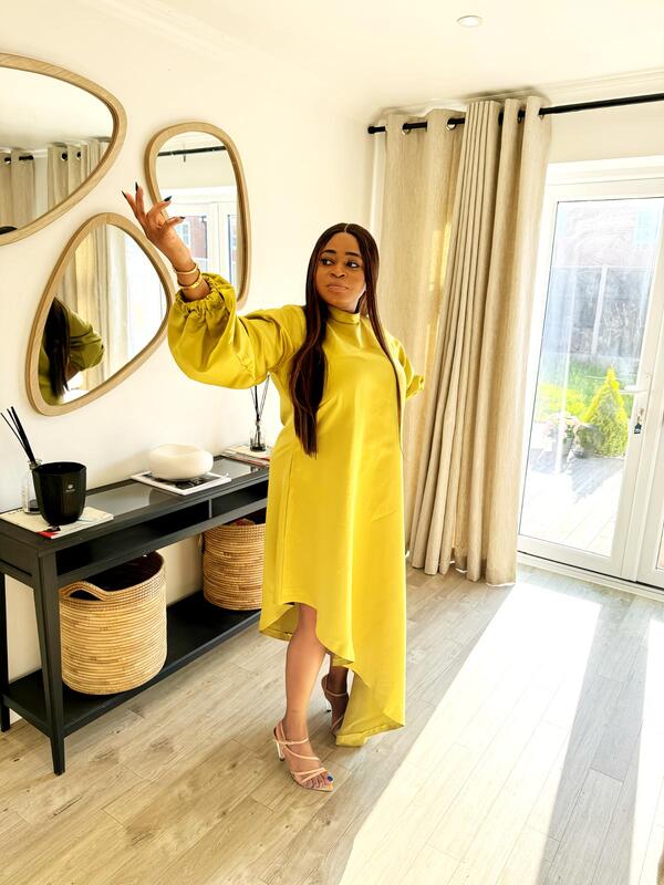 A woman wearing a bright yellow satin slanted dress with long puff sleeves and a high-low hemline, posing indoors near a mirror and window.
