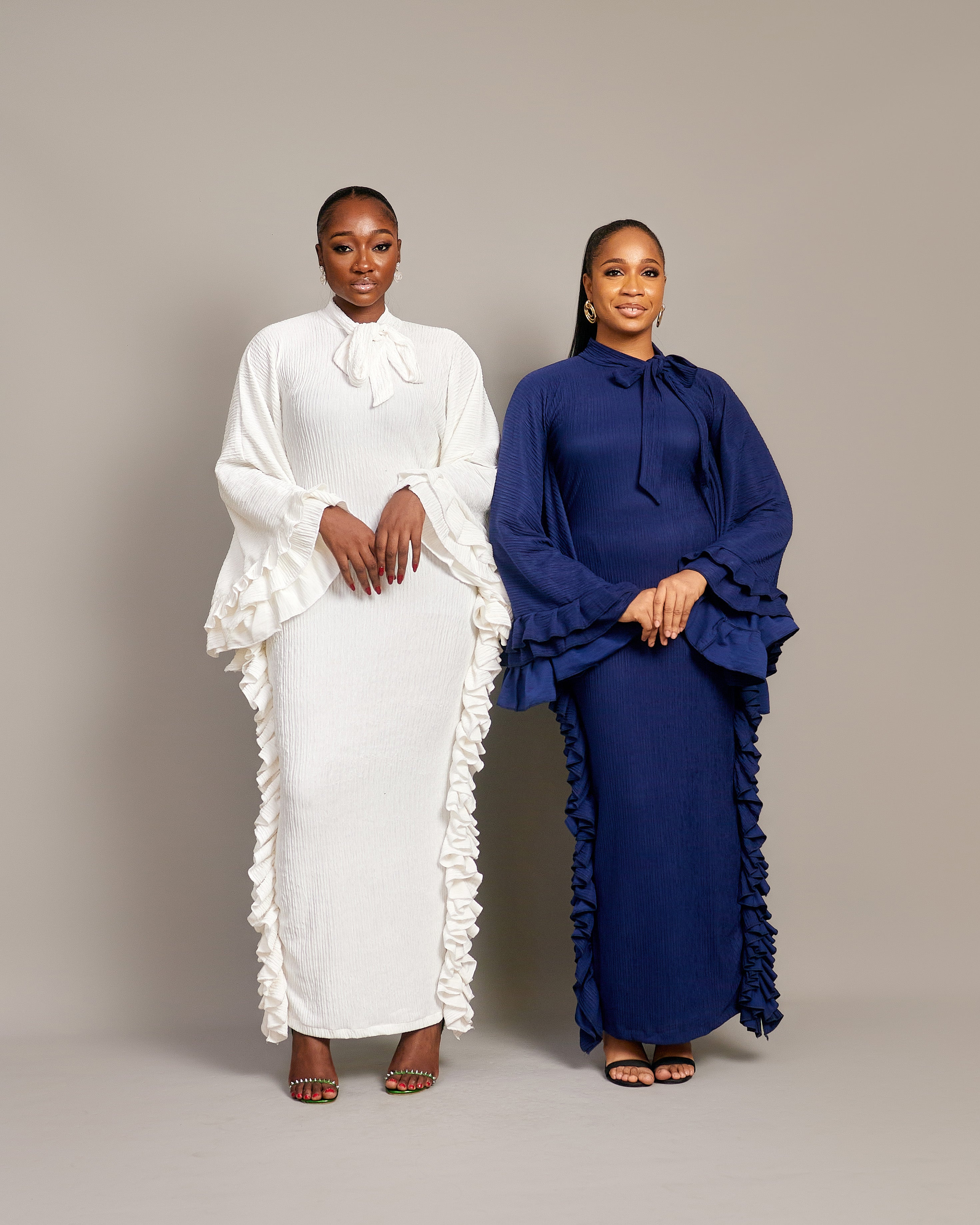 Two women wearing the Tella Dress: one in ivory, one in navy. Maxi gowns with ruffle sides, long sleeves, and bow-tie neckline.
